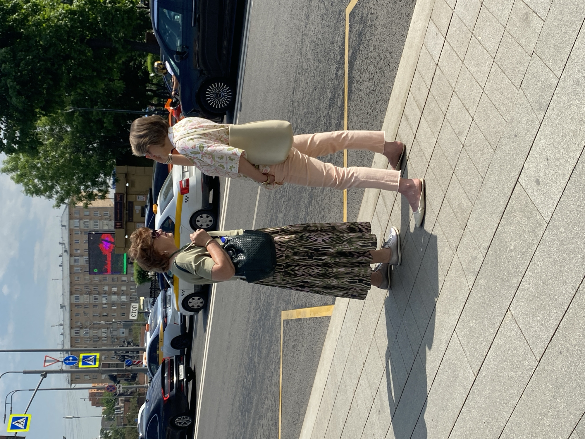 This image depicts a sunny urban street scene in Moscow, Russia, featuring two women standing on a tiled sidewalk next to a busy road. The strong sunlight creates clear, distinct shadows on the light grey pavement, indicating a bright day.
On the left side of the frame, a woman with short brown hair, wearing sunglasses, an olive green short-sleeved top, a long patterned skirt (green, brown, black), and grey sneakers, stands with a dark green woven shoulder bag. Slightly behind her and to the right, another woman with a lighter bob haircut wears a light floral blouse (white, green, pink), light peach trousers, and light-colored flat shoes, carrying a cream/beige shoulder bag. She is looking slightly to her right.
Adjacent to the sidewalk, a yellow line marks the edge of a road filled with vehicles. Several white and yellow taxis, characteristic of Russian public transport with their yellow stripe and checkerboard patterns, are visible, along with dark-colored sedans. One taxi's license plate, "M 837 OO 77," is discernible.
In the background, multi-story buildings line the street, with a large digital billboard displaying colorful abstract patterns in red, green, and blue on one of them. Lush green trees are also scattered throughout the background. Various street signs are visible, including blue square pedestrian crossing signs, a red triangular warning sign, a blue round sign indicating a right turn, and a white rectangular sign with a red border that clearly reads "СТОП" (STOP) in Cyrillic. Smaller informational signs bearing codes like "C271" and "0274" can also be seen.