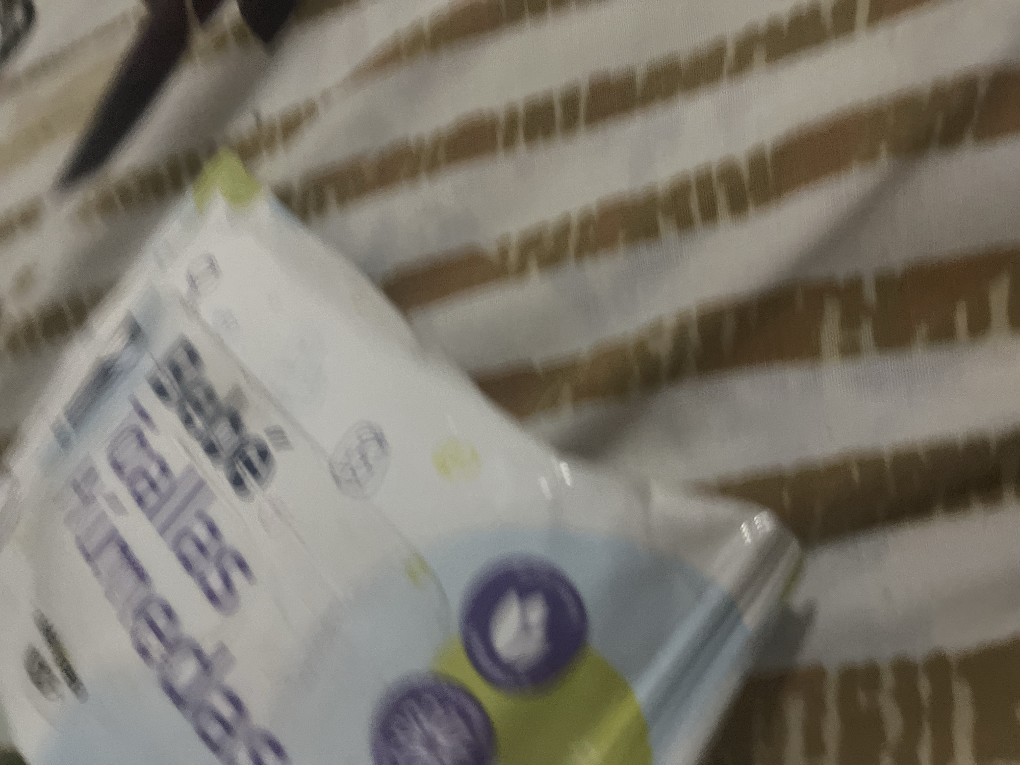 The image shows a close-up of a package of baby wipes. The text on the package is blurred but appears to be in Spanish. The package is light blue and white with some purple circles. The background is a blurry striped fabric, likely a bedsheet or blanket, in shades of white and brown. The image is taken indoors. There is no discernible information about the location in Santa Ana, Venezuela.