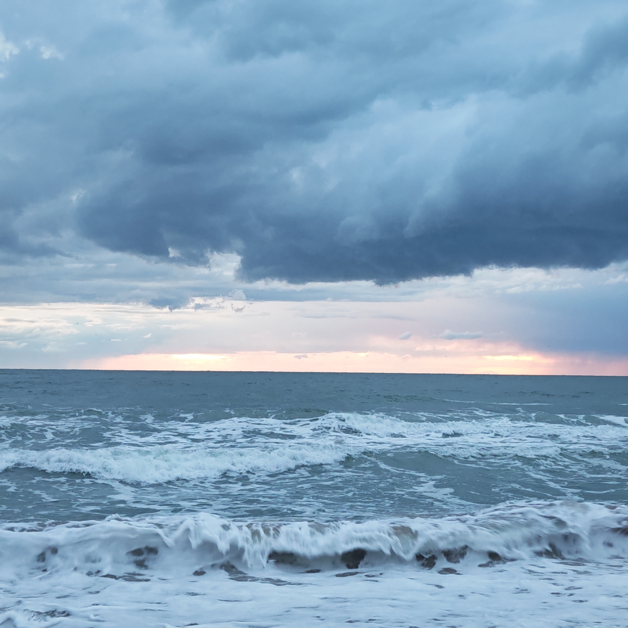 The image depicts a seascape under a cloudy sky, likely a beach scene. The foreground shows waves breaking on the shore, with white foam contrasting against the darker water. The sea extends to the horizon, where a faint glow of light is visible through a break in the clouds. Above the horizon, dark and heavy storm clouds dominate the sky.
There are no people visible in the scene.
Given the provided location information, this could be a beach scene in городское поселение Щелкино, Ukraine, a coastal town, though specific location confirmation requires visual landmarks.