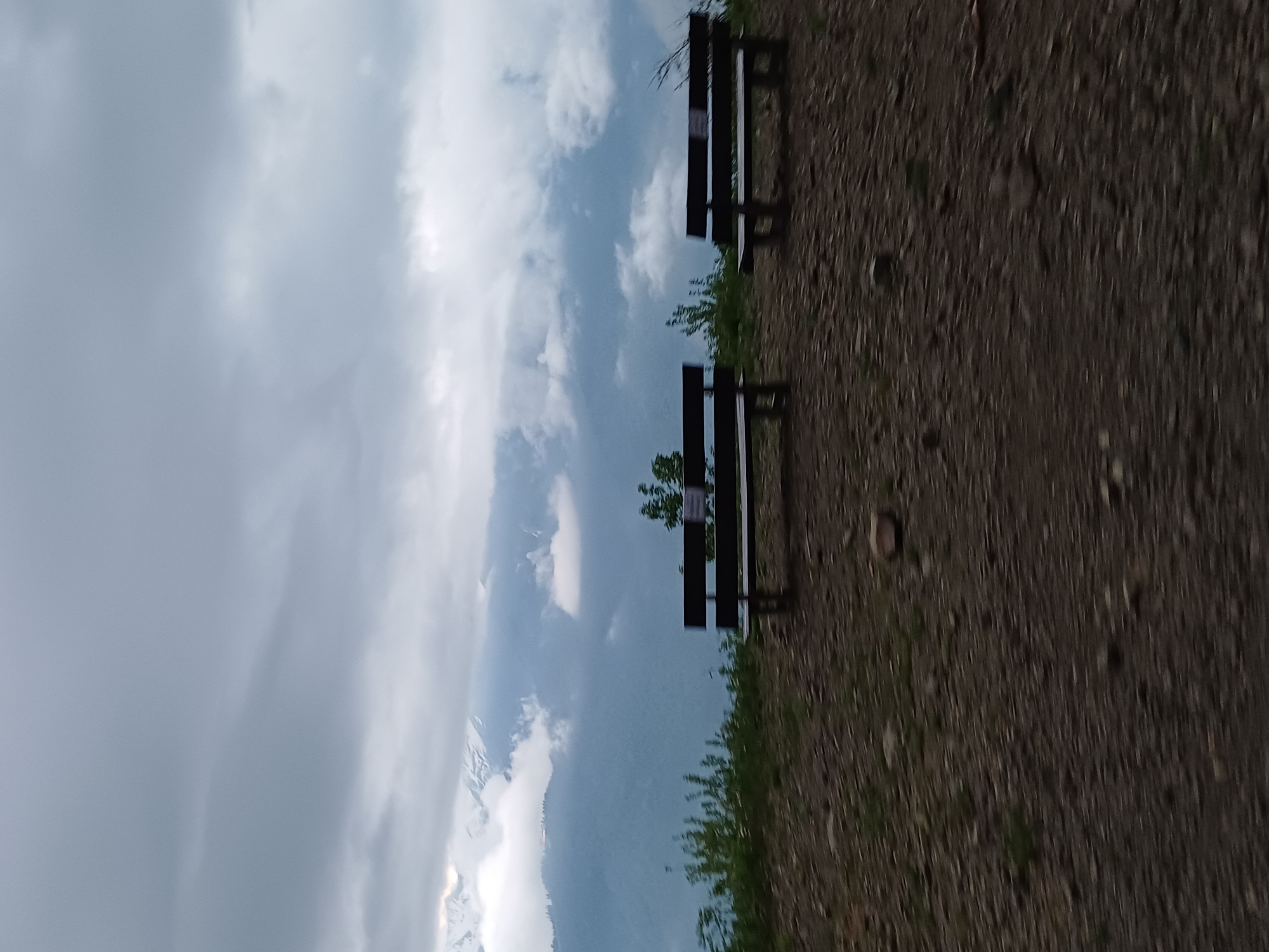 The image depicts a scenic mountain overlook in Estosadok, Russia, featuring two dark wooden park benches positioned on a gravelly path. The benches are oriented towards a vast landscape of layered, mist-shrouded mountains under a dramatic, cloudy sky. The foreground is dominated by the rough texture of the path, with scattered stones and sparse green vegetation. In the background, the mountains rise in successive planes, with some peaks appearing snow-capped, while others are obscured by soft, grey clouds. The overall atmosphere is serene and somewhat dramatic due to the overcast sky and the imposing mountain scenery. There are no people visible in the image.