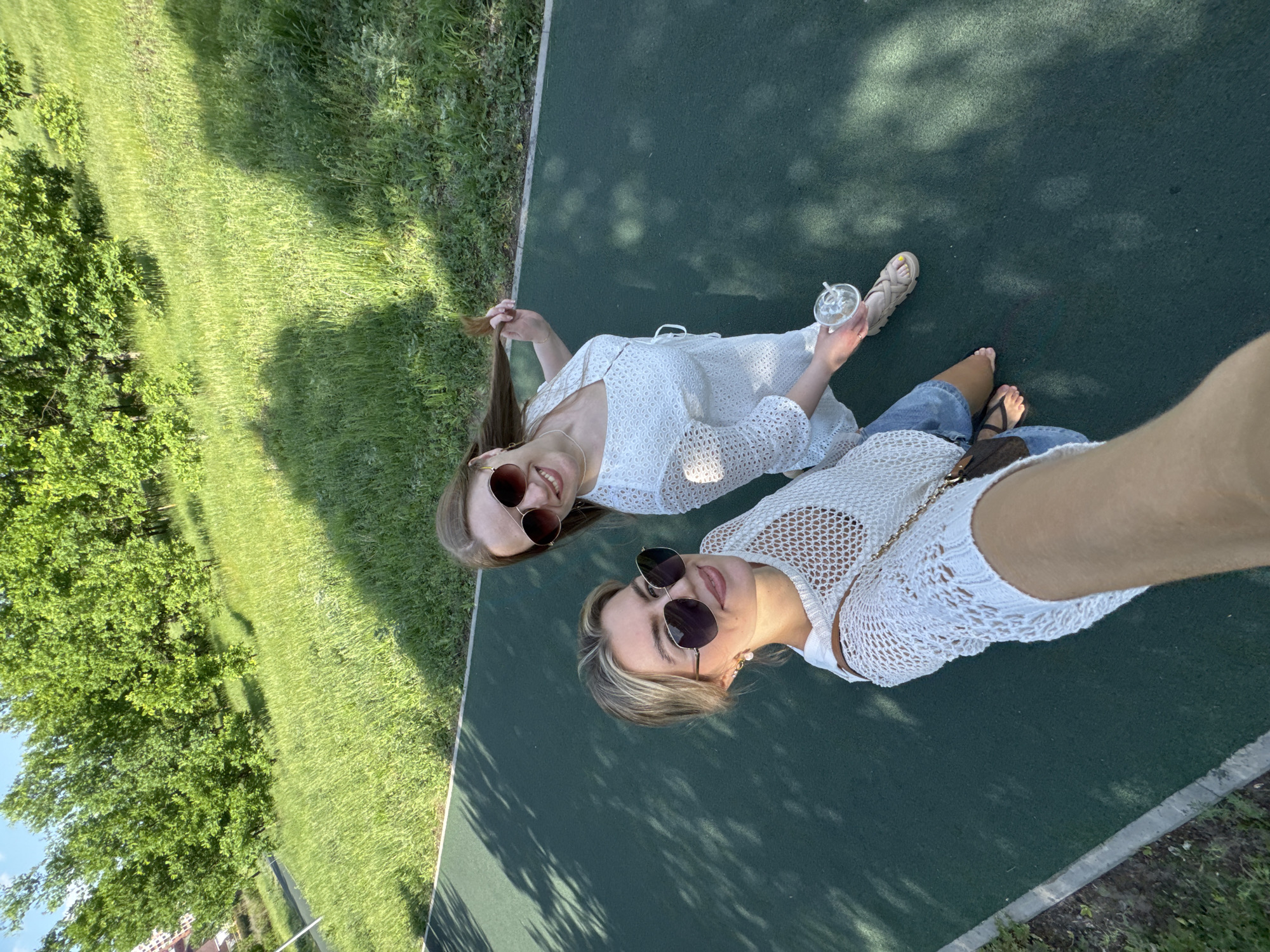 Two women are lying on a green surface, likely a tennis court or similar sports surface, with a grassy area to the left. The woman on the top is wearing a white eyelet dress, brown sunglasses, and holding a clear plastic cup with a straw. The other woman is wearing a white crochet top, blue denim shorts, and brown sunglasses. Both women are smiling, suggesting a pleasant experience. The location is Unknown, Unknown, but appears to be a public park or recreational area within a city.