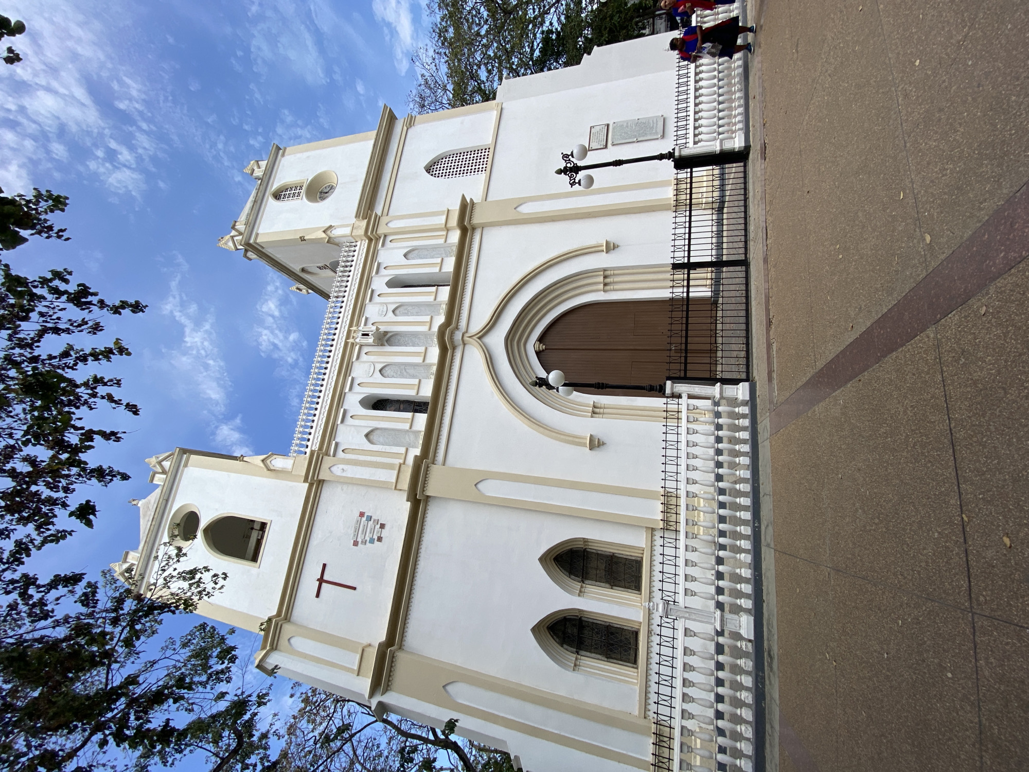The image shows the Catedral de Nuestra Señora de la Asunción in Porlamar, Venezuela. The cathedral is a white building with light beige accents, featuring a large arched wooden doorway and two towers. A black metal fence runs along the side of the building, with a white concrete railing visible behind it. A black lamppost stands in front of the building. The ground to the right is a tan-colored paved area with a reddish-brown stripe. In the upper right corner, a couple of people wearing red and blue clothing are standing near the edge of the frame. Trees with green foliage are visible on the left and above the building. The sky is blue with scattered white clouds. A red cross is on the building as well.
