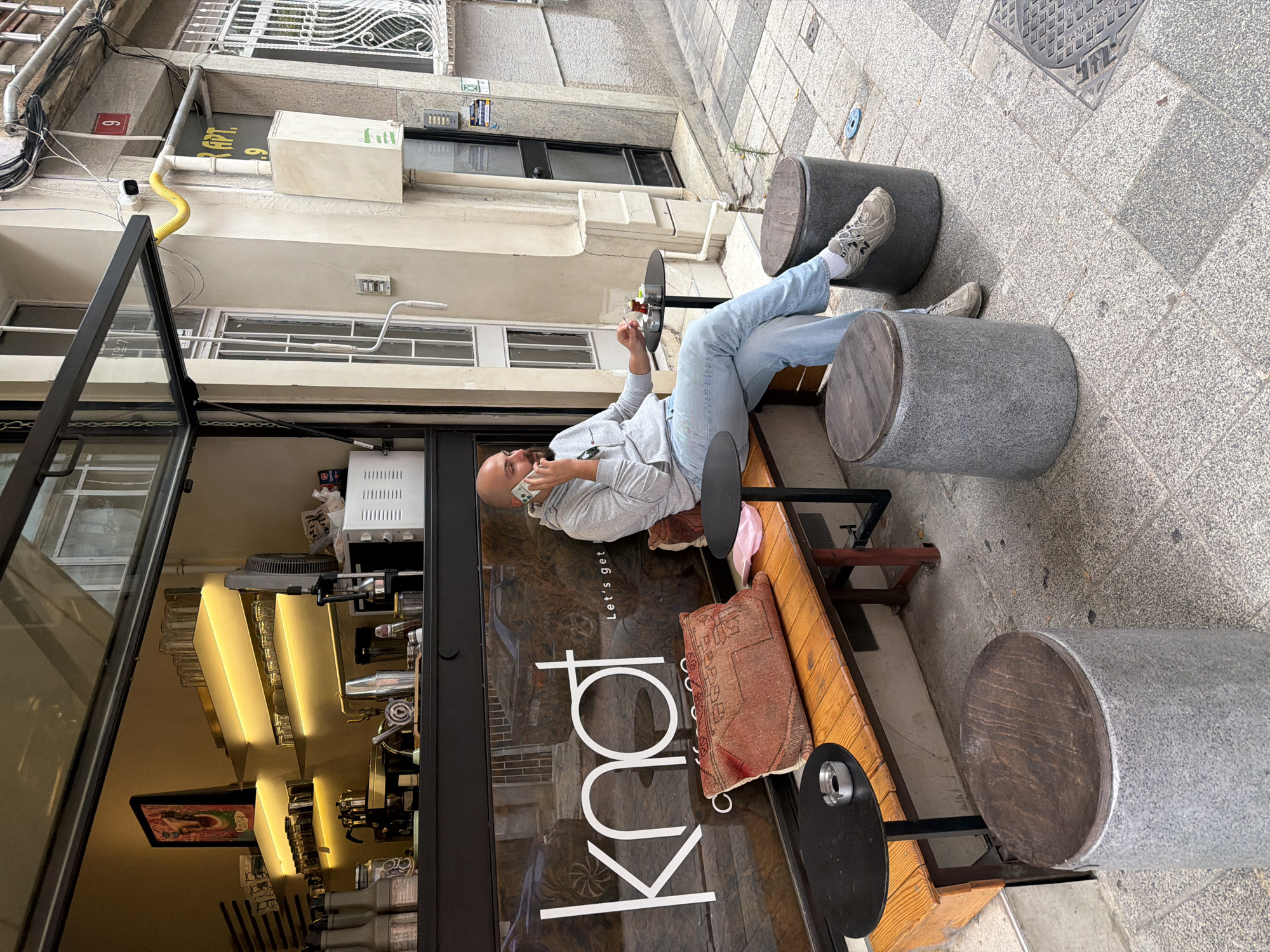 A bald, light-skinned man is seated on a wooden bench outside a cafe, talking on his mobile phone. He is wearing a grey hooded sweatshirt and light blue jeans, with grey sneakers and white socks. In front of him, on a small black table, are a glass of tea, an ashtray, and a pack of cigarettes. The bench has a red patterned cushion and a pink item on it.
The cafe has a large glass window with the white text "knot" and "Let's get..." visible. Inside the cafe, shelves are stocked with glasses and bottles, and a microwave is visible. Outside, there are two grey cylindrical stools with wooden tops. The cafe is located on a paved sidewalk.
The cafe appears to be located in Kadıköy, Turkey, given the details visible in the background, such as the building numbers and architectural style. A red sign with the number "9" is visible on the building next to the cafe. Above the cafe entrance, a sign reads "APT. 9". The street appears to be a commercial area with shops and cafes.