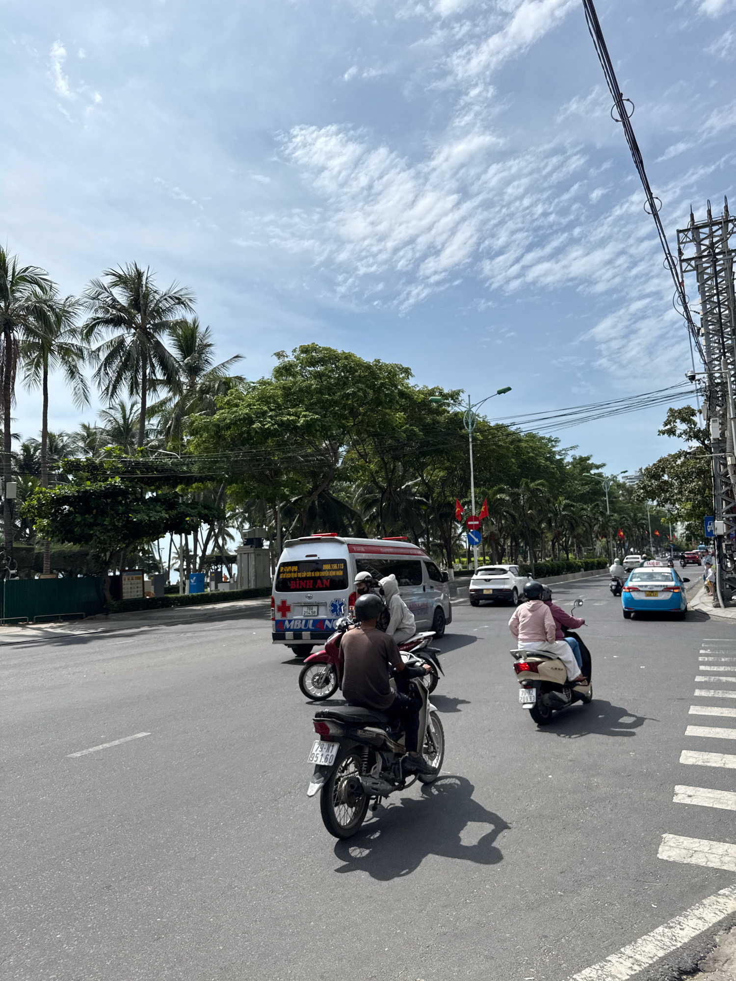 A street scene in Nha Trang, Vietnam, features a multi-lane road with various vehicles. In the foreground, two motorcycles are visible. The motorcycle on the left has a rider with a dark shirt and helmet. The motorcycle on the right is ridden by a person in a pink jacket and white pants, with a passenger also in white. In the middle ground, an ambulance is stopped, and a white car is moving past it. Further down the road, a light blue taxi is in motion. The background shows a dense line of palm trees and lush green foliage, with a cloudy sky above. Electrical poles and wires are present on the right side of the image.