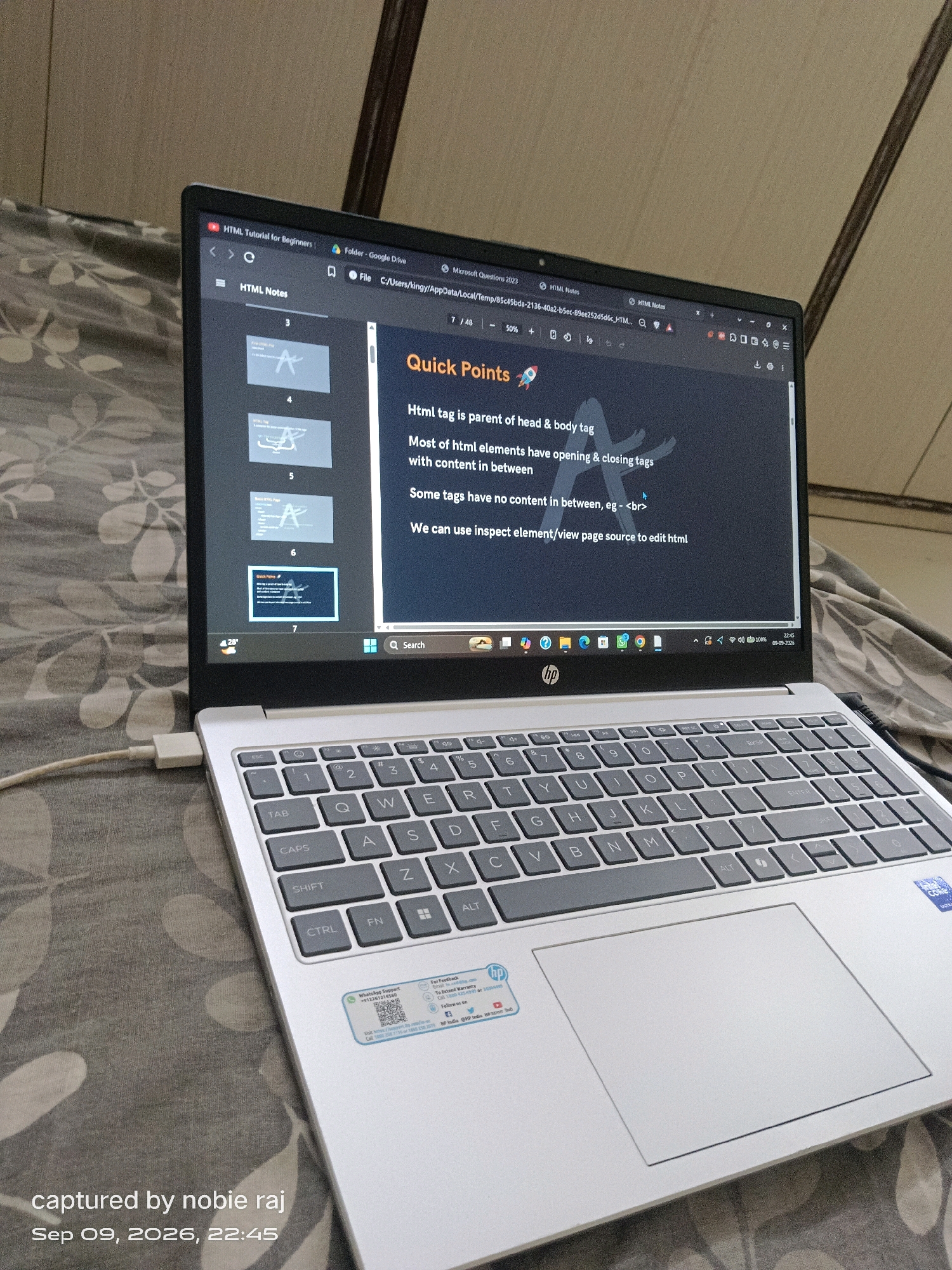 An HP laptop is open and placed on a bed with a patterned gray comforter. The laptop screen displays a presentation titled "HTML Tutorial for Beginners," with a slide showing "Quick Points" about HTML tags. The keyboard is visible, with a white power cord plugged into the left side of the laptop. The scene is dimly lit, suggesting it might be nighttime or an indoor setting. The bottom of the image contains a timestamp indicating "Sep 09, 2026, 22:45" and the text "captured by nobie raj." The overall impression is one of a person using a laptop for educational purposes in a relaxed, personal environment. There is no identifiable connection to Ganiyari, India, or any specific city.