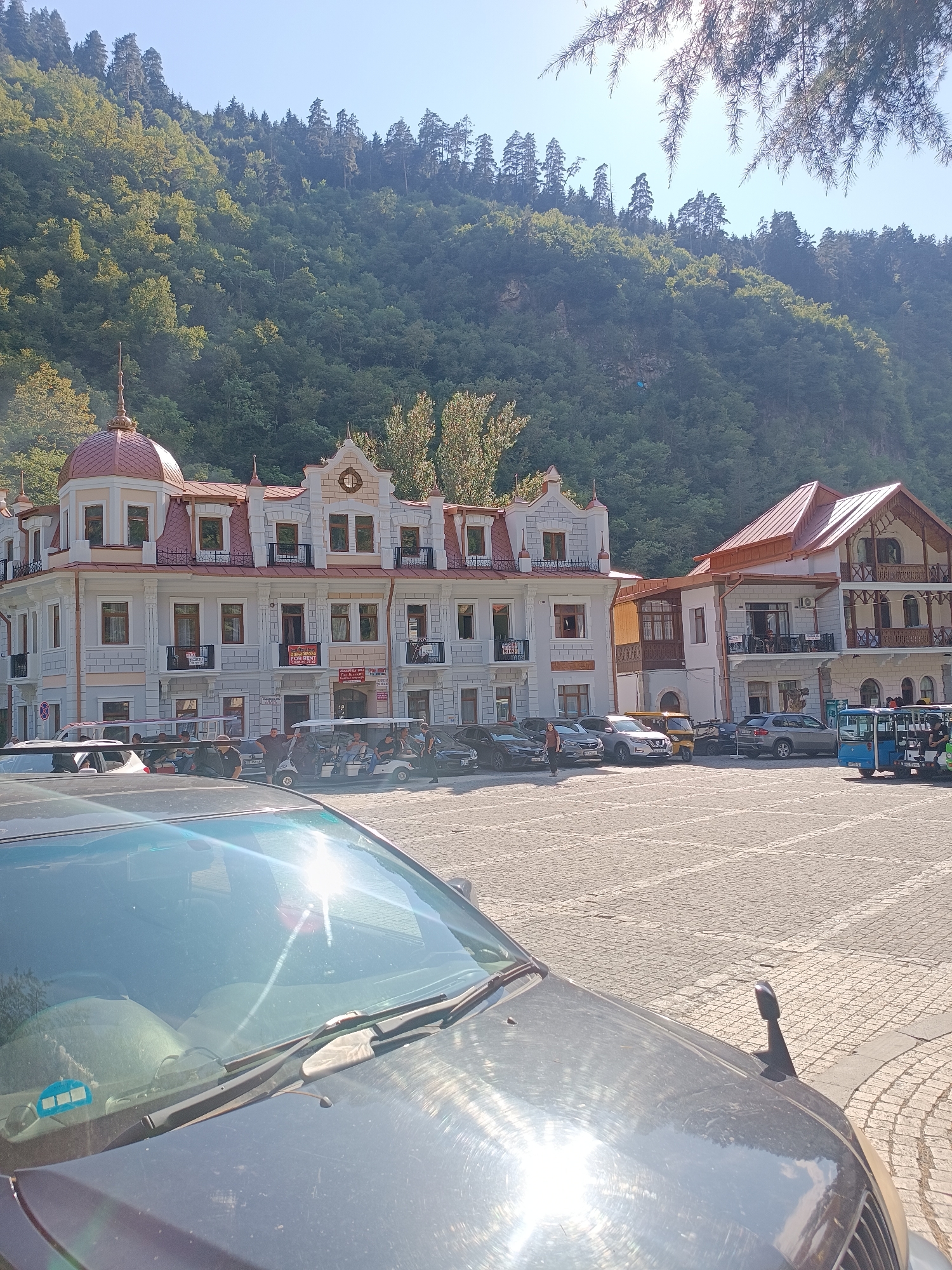 The image depicts a street scene in Borjomi, Georgia, on a sunny day. In the foreground, the hood and windshield of a dark-colored car are visible, with sun glare reflecting off the glass. The car is parked on a cobblestone street.
Behind the car, several buildings are prominent. The largest building is a multi-story structure with a white facade, ornate architectural details, and a copper-colored domed roof. It has multiple windows with balconies. Adjacent to this building is another, slightly taller structure with a similar facade and a pitched, copper-colored roof. Both buildings appear to be commercial or residential properties.
Parked in the street are various vehicles, including several cars, a blue bus, and what appear to be golf carts or small utility vehicles. A group of people, predominantly male, are standing and gathered around some of these vehicles. One person is visible walking away from the camera on the left side of the street.
In the background, a steep, tree-covered mountain slope rises behind the buildings, with a clear blue sky above. Pine trees are visible in the upper right corner, obscuring a portion of the sky and mountain. The overall scene suggests a town square or a street in a town situated within a mountainous, forested area.
