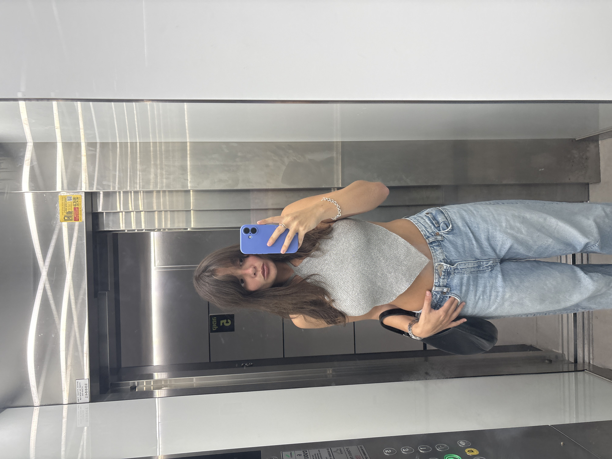 A young woman with long, dark brown hair and bangs stands in an elevator, taking a selfie in a mirrored wall. She is wearing a gray, halter-neck crop top and light-wash, baggy jeans. She holds a purple smartphone in her right hand and a black shoulder bag in her left. The elevator interior is metallic with a panel of buttons visible on the left side, and a sticker with Hebrew text is affixed to the upper right wall. The elevator doors are closed. The scene is likely inside a residential or commercial building in Tel-Aviv, Israel, given the visible Hebrew text and common elevator design.