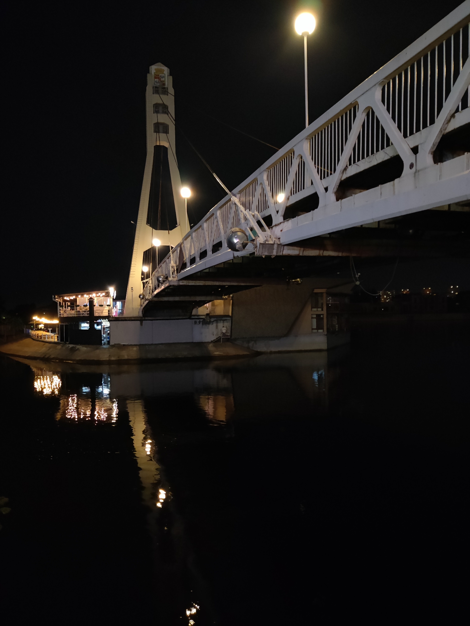 This is a nighttime photograph of a bridge spanning a body of water. The bridge is illuminated by artificial lights, with streetlights visible along its walkway and the structure itself casting a white glow. The water below reflects these lights, creating shimmering patterns.
On the left bank of the water, a building with outdoor seating, possibly a restaurant or cafe, is visible. It is also lit up with string lights. A tall, white tower, part of the bridge's suspension system, stands prominently behind the building and bridge. The sky is dark, indicating it is night.
There is no apparent activity or event taking place, other than the general illumination of the scene. Notable details include the reflections in the water, the architectural design of the bridge and tower, and the inviting lights of the building. The location is unknown, and the weather appears calm. No visible text is present in the image.