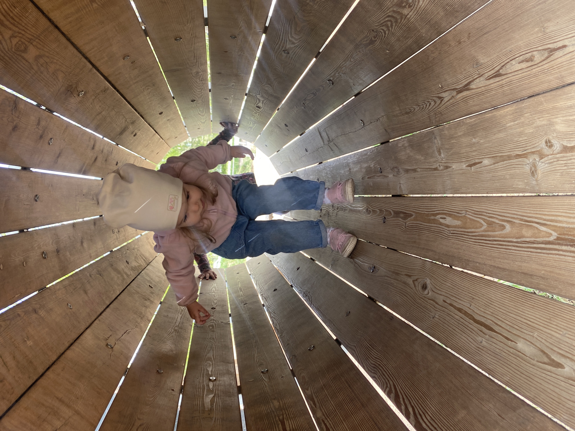 A young child is crawling through a wooden tunnel structure in Perm, Russia. The tunnel is constructed from wooden planks with gaps between them, allowing light to stream in. The child wears a light pink sweatshirt, blue jeans, a beige hat, and pink shoes. The interior of the tunnel creates a perspective with converging lines leading toward an exit visible in the distance. The wood grain is visible on each plank, revealing variations in color and texture. Metal fasteners are visible, securing the planks together. The child is positioned centrally within the tunnel, appearing to crawl towards the opposite opening.