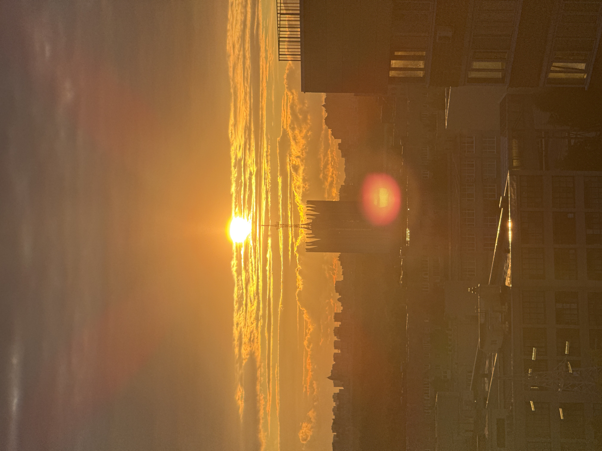 This image depicts a city skyline at sunset, bathed in a warm, golden light. The sun, a bright orb, is partially obscured by striated clouds, casting long shadows and highlighting the architectural features of the buildings. A prominent, spire-like structure with gothic-inspired detailing stands out against the horizon, likely a significant landmark. In the foreground, modern buildings with visible windows and balconies occupy the lower portion of the frame, their surfaces reflecting the intense sunlight. The overall atmosphere is one of tranquility and urban grandeur, with the natural phenomenon of sunset juxtaposed against the man-made environment. The location is identified as Unknown, Russia.