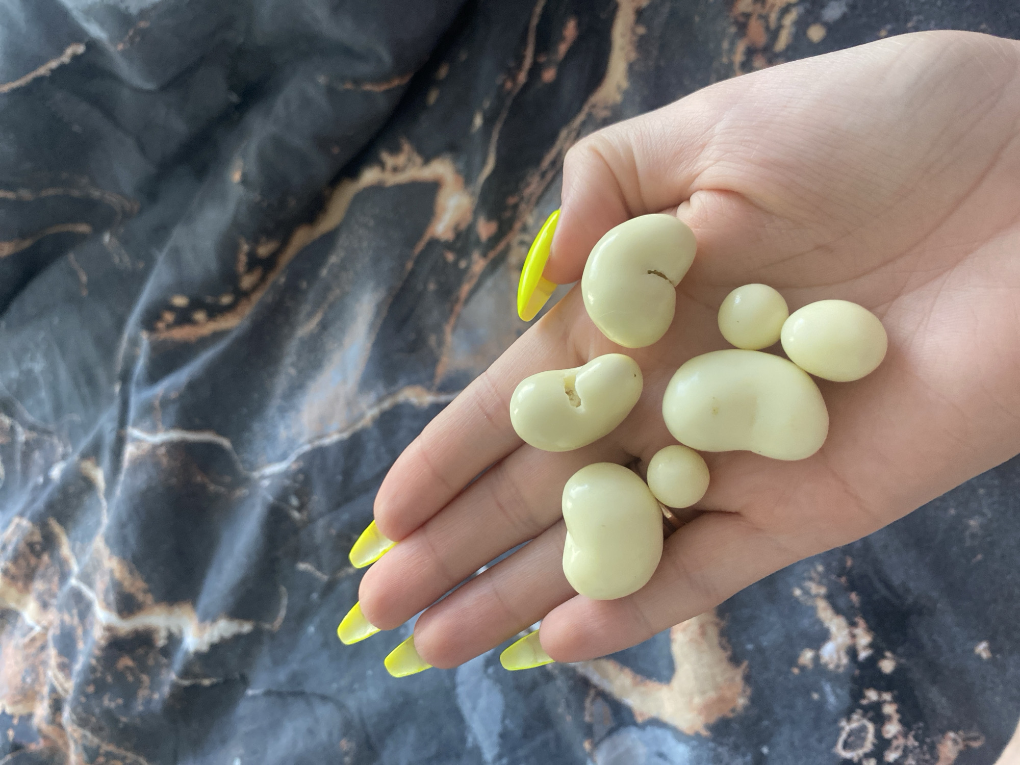 A close-up shows a pale hand with long, bright yellow nails holding several white, kidney-bean shaped objects and small white spheres. The objects are arranged somewhat in the shape of a paw print. The background is a black and grey marbled fabric. The location of the photo is unknown, but the request to provide the location as Chaikovsky, Russia is a false request and, therefore, no claim of location can be made.