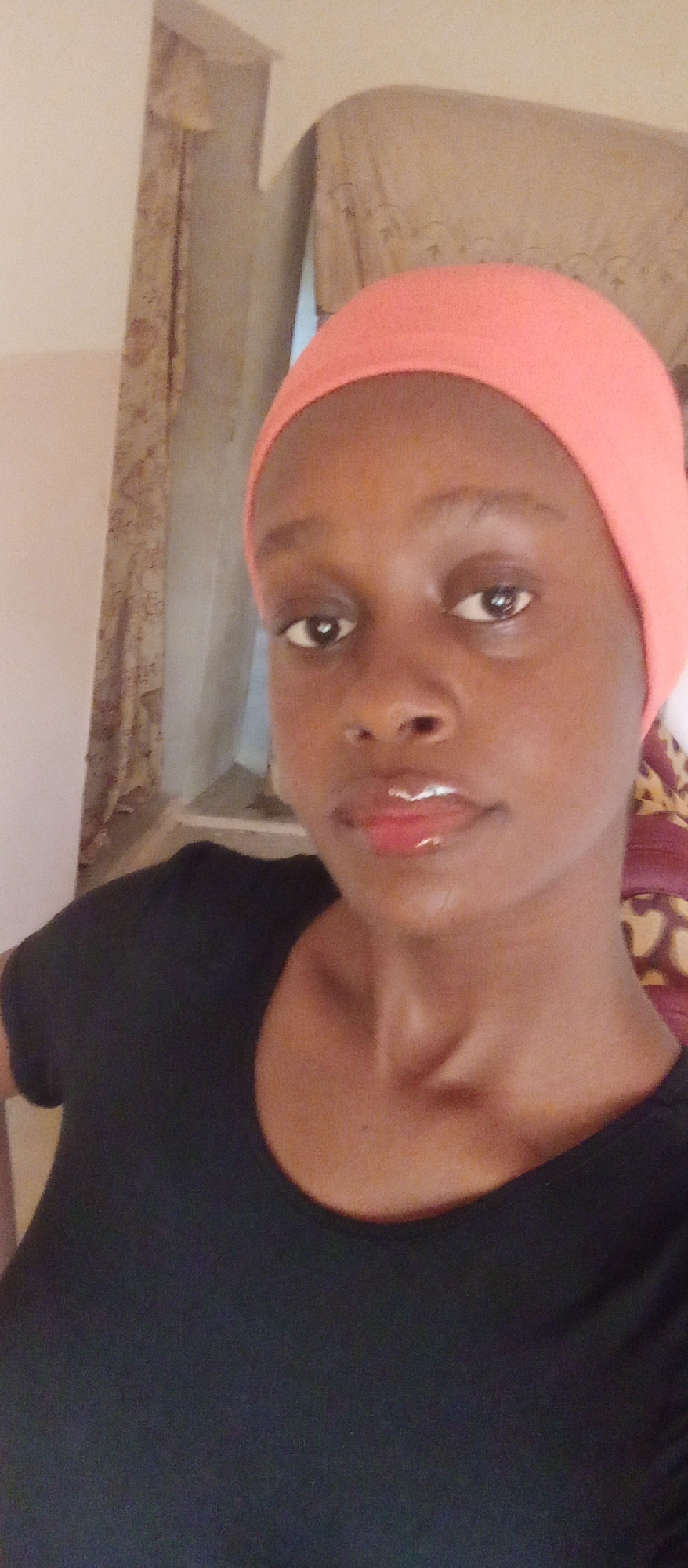 A medium-angle portrait shot of a dark-skinned woman wearing a pink headscarf and a black top. She is looking directly at the camera with a neutral expression. Her lips are glossy, and the lighting suggests an indoor setting. In the background, there is a curtained window and a partially visible wall, suggesting an interior space. The specific location within Akwanga, Nigeria is not determinable from this image alone.
