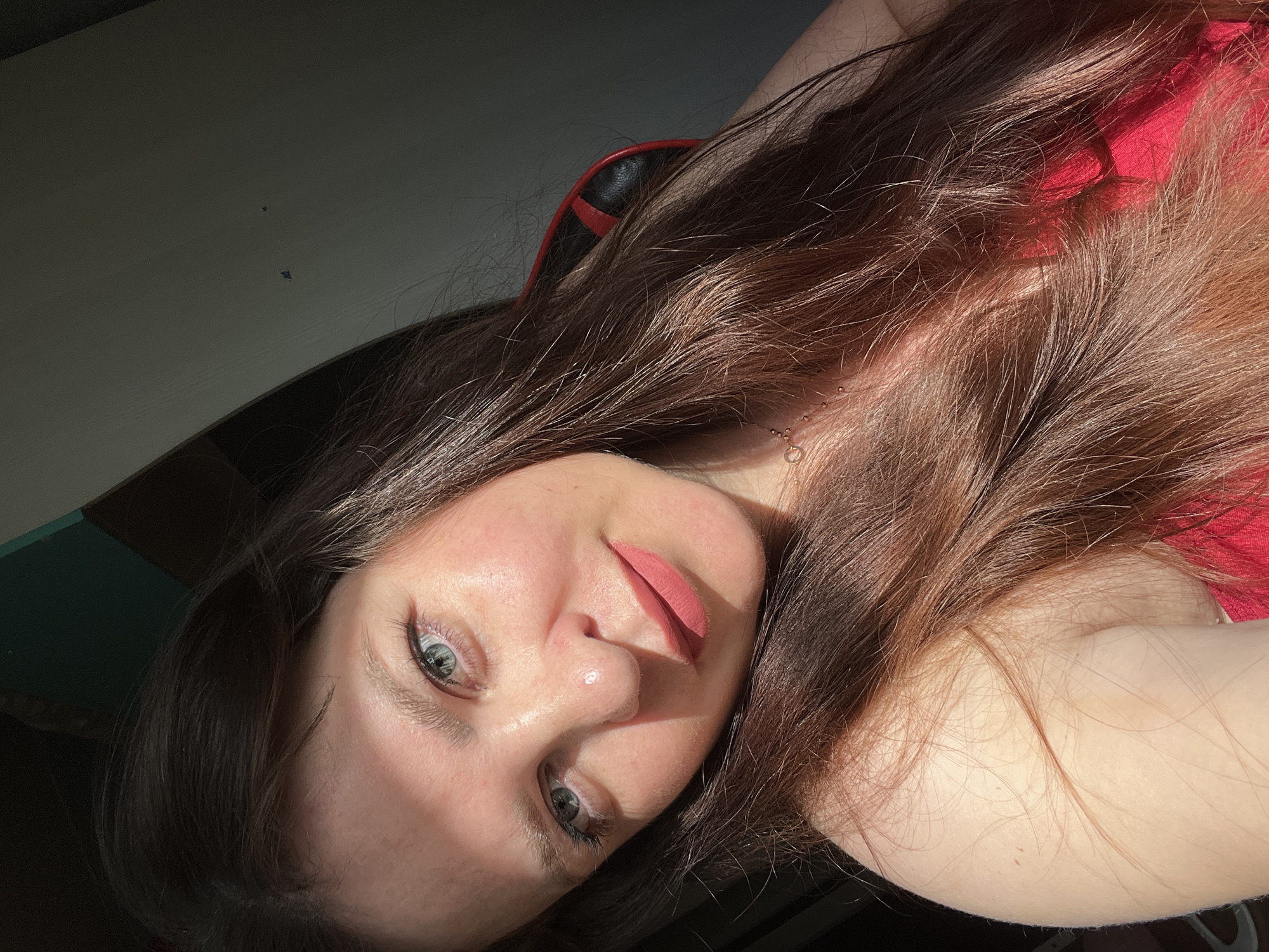 The image shows a close-up, selfie-style shot of a young woman with long, brown hair and blue eyes. She is wearing a pink lipstick and a red top. Sunlight is casting a shadow across her face. Her hair is partly covering her left shoulder and neck. The background is dark, with some hints of a wall and possibly furniture, but it is not detailed enough to determine a specific location within Noviy, Russia.