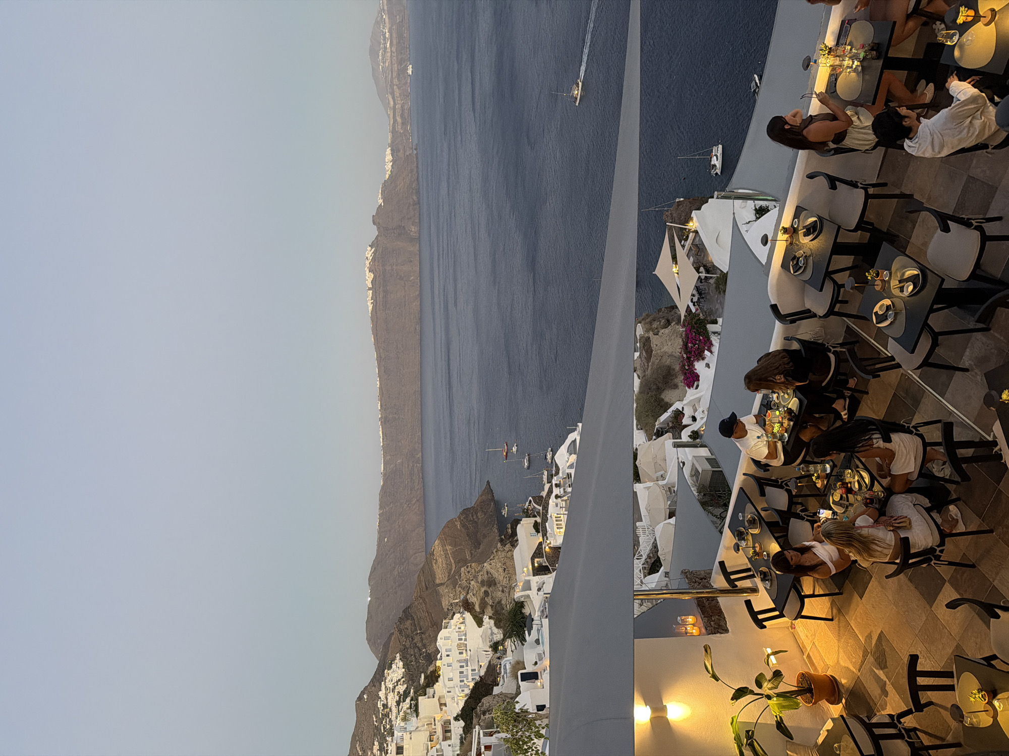The image captures a view of Oia, Santorini, Greece, during twilight. The upper portion of the image displays the caldera with whitewashed buildings clinging to the cliffs and the deep blue Aegean Sea. Several boats are visible on the water. The lower portion of the image shows an outdoor restaurant terrace. People are seated at various tables, some dining, others conversing. The atmosphere is dimly lit, with warm light emanating from lamps on the tables and wall sconces. The tables are set with plates, glasses, and small decorative items. A potted plant adds a touch of greenery to the scene. The architecture of the buildings, with their characteristic white walls and blue accents, is typical of Santorini.