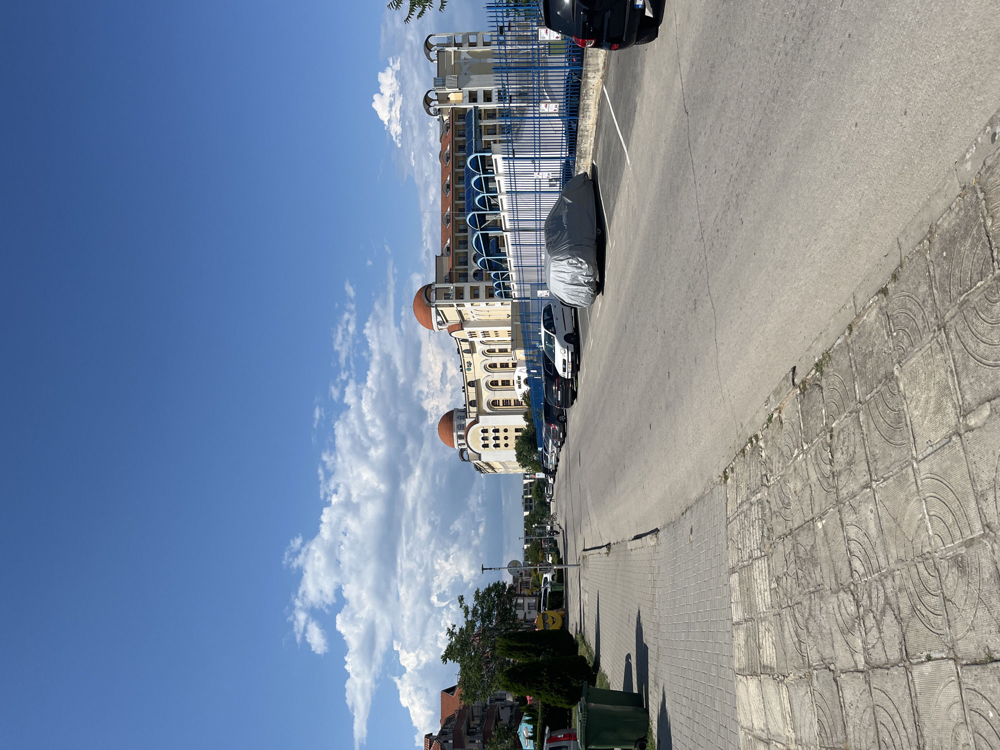 The image depicts a street scene in the city of Obzor, Bulgaria, on a bright, sunny day with scattered clouds. The perspective is from the sidewalk, looking down a paved road lined with parked cars. On the left side of the road, there is a sidewalk with decorative patterned paving slabs, followed by a row of green trash bins and some cypress trees. Beyond the trees, residential buildings with tiled roofs are visible.
On the right side of the road, a large, light-colored building with red-tiled domes and an arched colonnade is prominent. It appears to be a hotel or resort complex, with a blue metal fence along its perimeter. Several cars are parked in a designated lot in front of this building, one of which is covered with a gray tarp. The road itself is asphalt, with painted white lines indicating parking spaces. In the distance, the road curves slightly, and more buildings and trees can be seen. The sky is a vibrant blue with fluffy white clouds. No people are clearly visible in the image.
