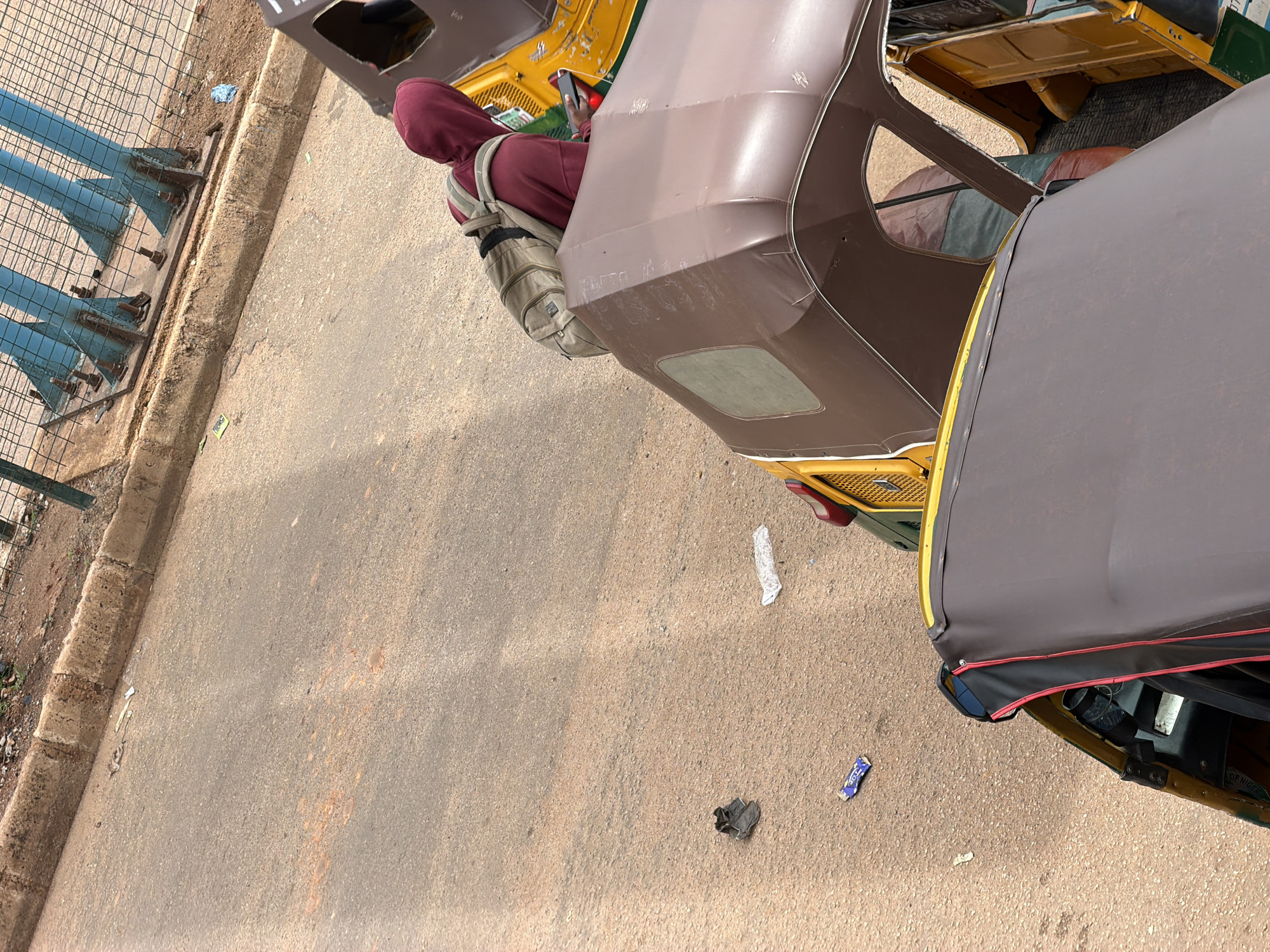 In Bukuru, Nigeria, the image captures a street scene with several auto-rickshaws. A person wearing a maroon hoodie and a backpack is visible, leaning over the canopy of one of the vehicles. The auto-rickshaws, common in urban Nigeria, are parked on a dirt road. A concrete curb with some debris lines the edge of the road, and a metal fence with blue structural supports is visible in the background. Scattered on the road are various pieces of litter, including a pair of dark shoes and some plastic wrappers. The lighting suggests it is daytime.