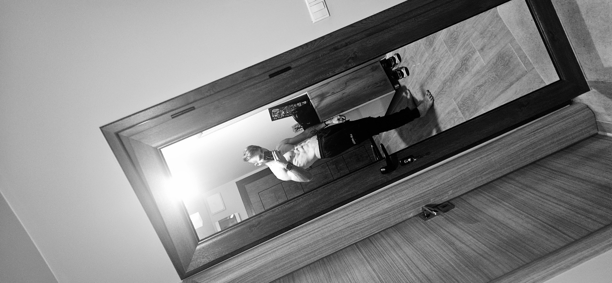 A black and white photograph depicts a mirror reflecting a man taking a selfie. The man, who is light-skinned, is wearing dark pants. His torso is bare, revealing abdominal muscles. He holds a device, presumably a phone, up to take the picture. The reflection also shows what appears to be a dresser or cabinet with a few items on top. His bare feet are visible. The mirror has a dark frame and is positioned at an angle. The floor is tiled. The location appears to be a room. The city or country cannot be determined from the image.