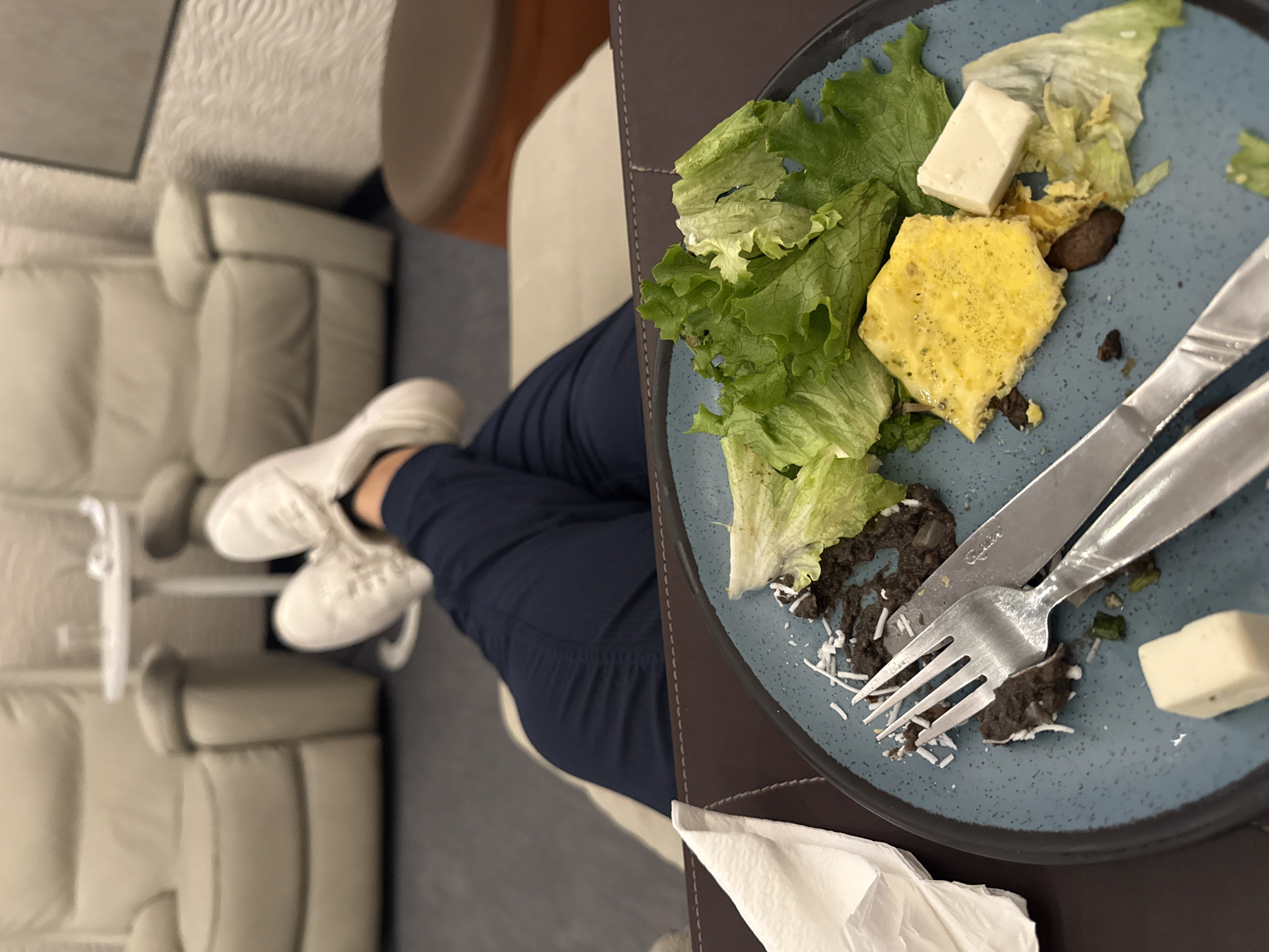 A person with white sneakers and blue pants sits on an ivory-colored couch and eats a salad consisting of lettuce, cheese, and an egg patty on a blue plate with silverware. A white napkin is placed below the plate on a brown surface. The location is Mexico City, Mexico.