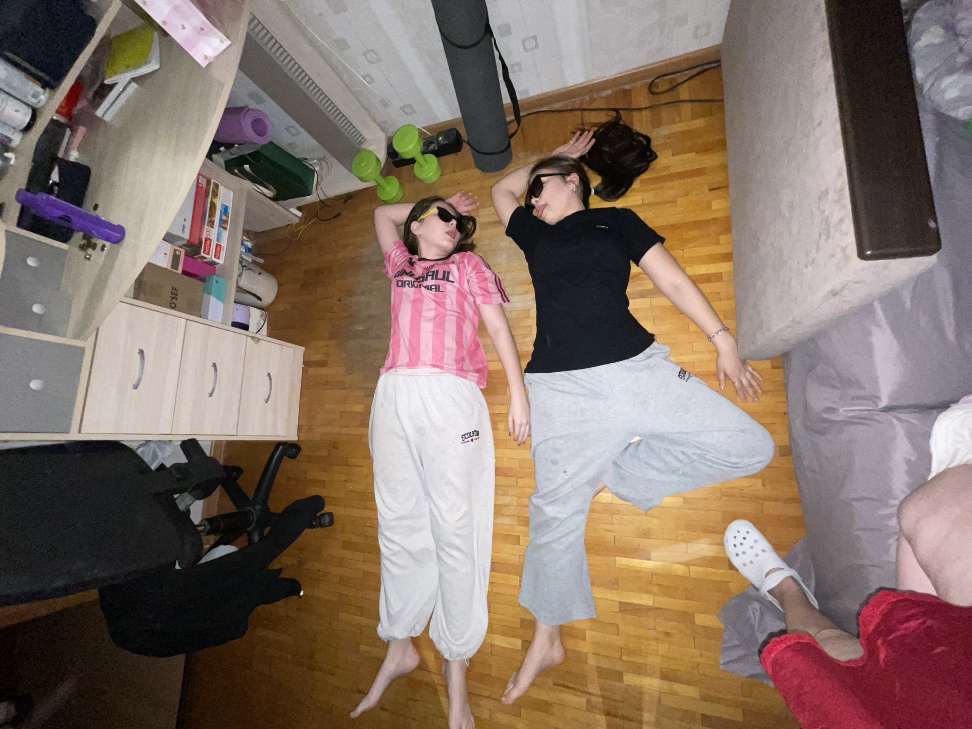 An overhead shot shows two young women lying on a light-colored wooden floor in an indoor room, identified as Murino, Russia. Both subjects wear dark sunglasses and appear relaxed or playfully posing; the woman on the left has her tongue slightly out.
The woman on the left is dressed in a pink and white striped short-sleeved shirt with the word "ORIGINAL" visible on it, and light-colored sweatpants that show the text "SCULPT". The woman on the right wears a black short-sleeved shirt and grey sweatpants. Both are barefoot. Another person's leg, wearing a white Croc-style shoe and red clothing, is partially visible in the bottom right corner of the frame, near the edge of a bed or mattress.
The setting appears to be a bedroom or a casual living space. To the left, a multi-tiered shelving unit is visible, containing various items such as books, boxes (one labeled "O'SEF"), and a purple yoga mat. A black office chair is positioned beneath a desk in the lower-left. Near the women's heads, a pair of green dumbbells and a rolled-up dark grey yoga mat are placed on the wooden floor. The overall scene suggests a relaxed and lived-in environment. No specific time of day or weather conditions can be discerned from the image.