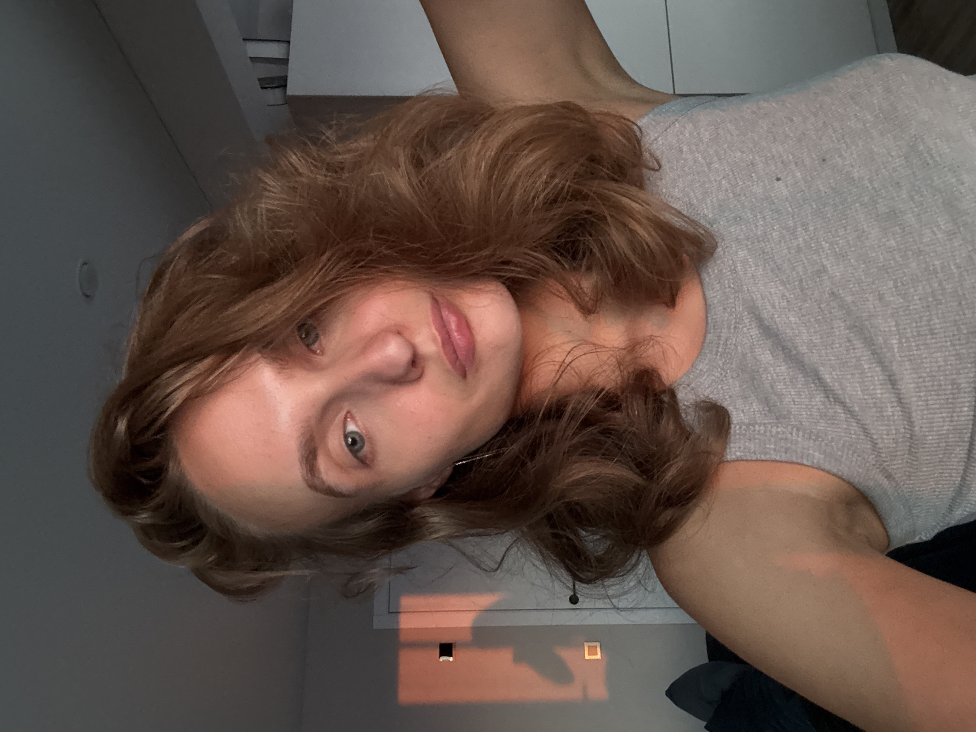 A young woman with auburn, wavy hair is looking into the camera. She is wearing a grey ribbed tank top. The lighting suggests it is either sunrise or sunset, with warm light illuminating her face and arm. A rectangular shadow, cast by a window, is visible on the wall behind her. The background includes a closed door, a section of a closet, and a dark couch with pillows. The overall impression is of a casual, indoor setting. The city of Подшивалово, Russia, is not depicted in this image.