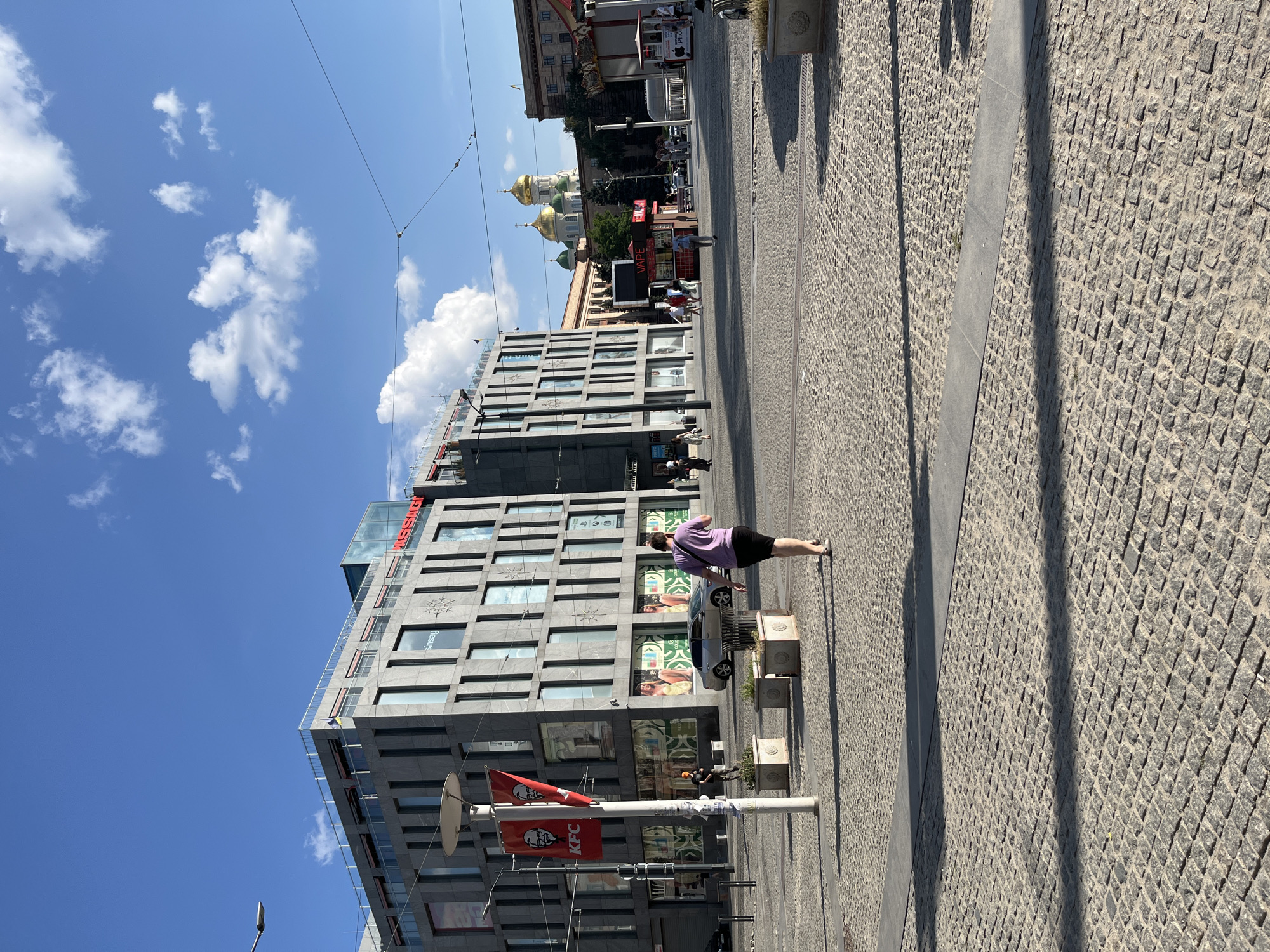 The image depicts a wide, paved public square in Dnipro, Ukraine, under a bright blue sky with scattered clouds. In the foreground, a person in a purple shirt and black shorts walks away from the camera across the cobblestone pavement. To the left, a modern, grey, multi-story building dominates the scene. It features large windows and signage, including a red KFC banner on a pole with the recognizable Colonel Sanders logo.
Further into the square, various kiosks and smaller buildings are visible. One kiosk is labeled "VAPE." In the background, a prominent Orthodox church with golden domes stands out against the sky, suggesting a historical or religious significance to the area. Several other buildings and people populate the square, some walking, others standing near the kiosks. The overall atmosphere is one of a bustling urban environment on a sunny day.