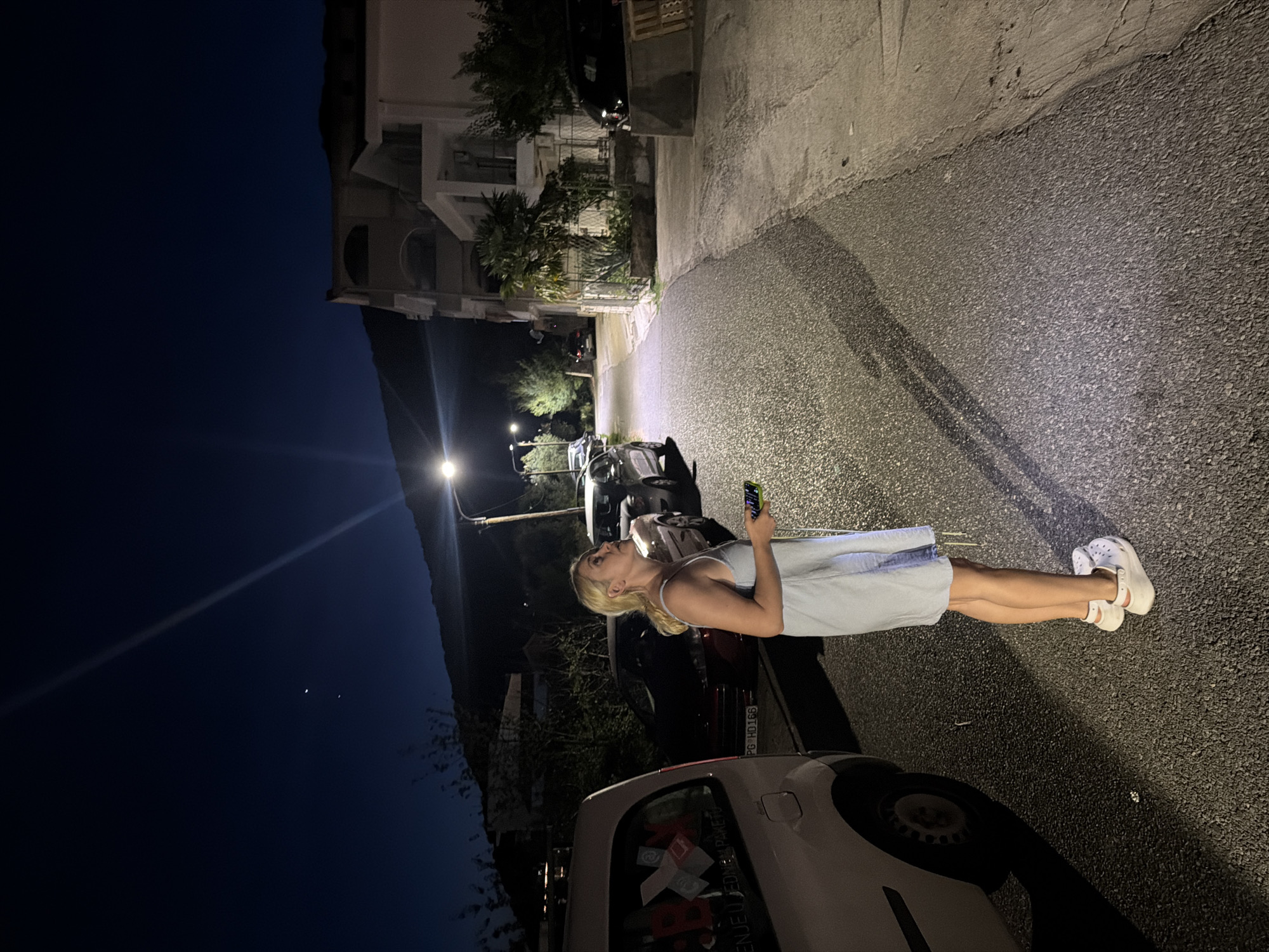 A young woman with blonde hair stands on a paved road at night in Petrovac na Moru, Montenegro. She is wearing a light blue sleeveless dress and white Crocs. The woman is holding a green smartphone in her right hand and looking up towards the night sky. In the sky, two celestial bodies are visible, possibly the moon and a planet. A street light illuminates the scene from the right, casting long shadows. Several parked cars are visible on the left and right sides of the road. A multi-story building is situated on the right side of the road, and a tree with palm-like fronds grows in front of it. In the background, a mountain range is silhouetted against the dark sky. The road surface is asphalt, and a concrete curb runs along the right side of the street. A white van is parked on the far left of the image, partially obscuring the view of the woman and the road.