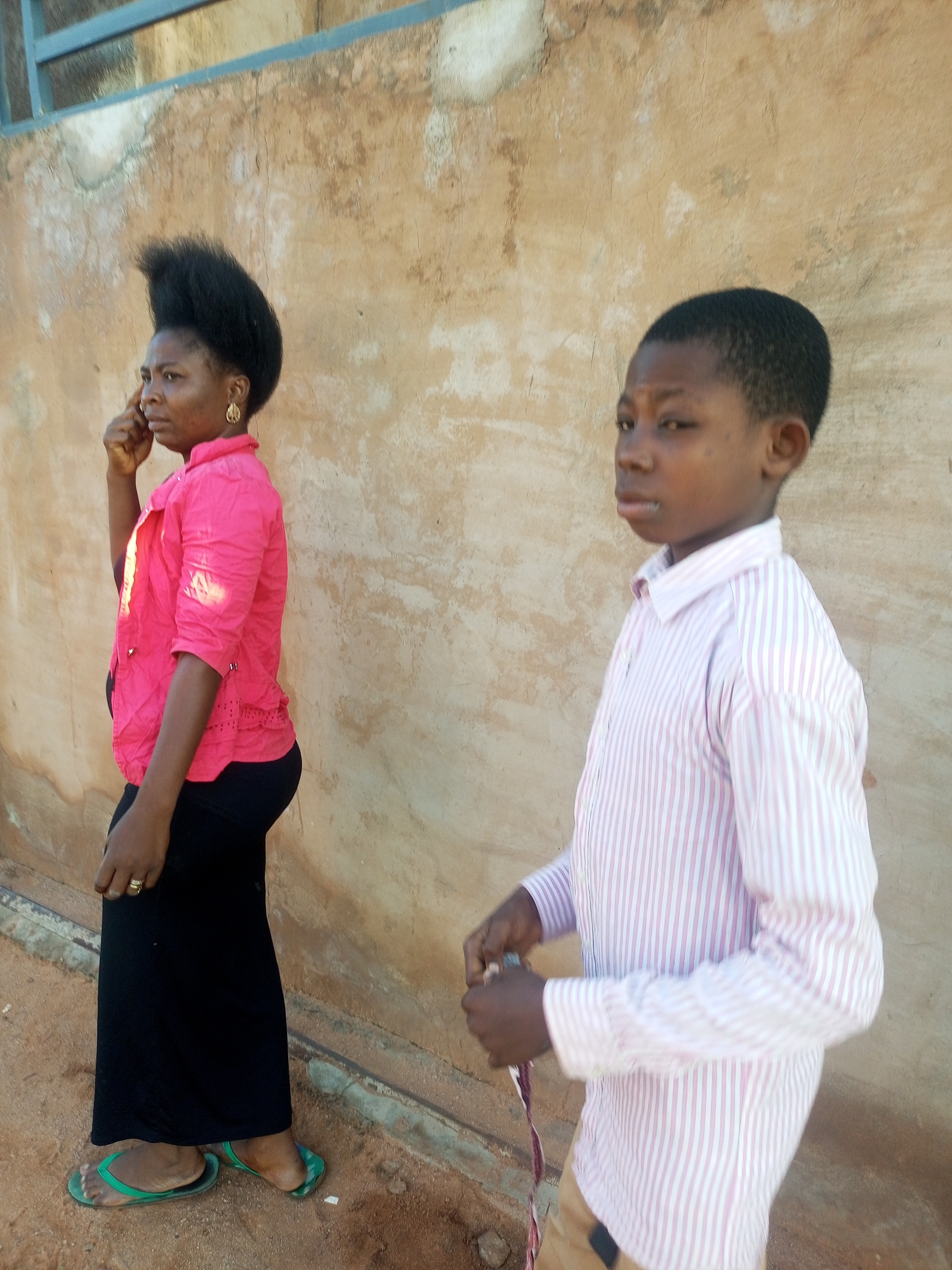 An outdoor, eye-level medium shot captures a woman and a young boy standing in profile against a textured, light brown wall. The woman, on the left, wears a bright pink, three-quarter sleeve button-up shirt with cut-out details on the hem and a long, black skirt. Her dark hair is styled upward in a voluminous manner, and she wears gold hoop earrings. She is holding a finger to her right ear as if speaking on a phone. A gold ring is visible on her left hand. She wears green flip-flops. The boy, on the right, is dressed in a pink and white striped button-up shirt and tan pants. He is holding a piece of rope or cord in his hands. The ground appears to be dirt. The lighting suggests it is daytime, with shadows cast on the wall and ground.