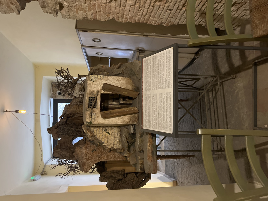 The image shows a model of a ruined building, located in a narrow, dimly lit hallway in Naples, Italy, at the coordinates 40.852 latitude, 14.257 longitude. The model is made of plaster, wood, and other materials and is painted in realistic colors. It depicts a small, single-story building with a crumbling facade and a doorway that is partially open. The model is supported by a wooden frame, and a sign with an inscription in Italian is placed on a table next to it. The sign appears to provide information about the model. There are two chairs in the hallway, one to the left of the model and another to the right. The chairs appear to be made of wood, and they are painted in a light brown color. The floor of the hallway is made of tile, and there is a light fixture on the ceiling. The hallway has white plastered walls, one of which is part of a larger structure made of exposed red brick. The hallway walls have a faded greenish hue on the lower portions. A large white cabinet is positioned to the right of the model. The cabinet is divided into two halves by a thin vertical dividing line and has a metallic finish. The cabinet is partially obscured by the wall on the right side. The model is the primary subject of the image. The scene is dimly lit, with shadows being cast on the floor. The image is taken at eye-level.