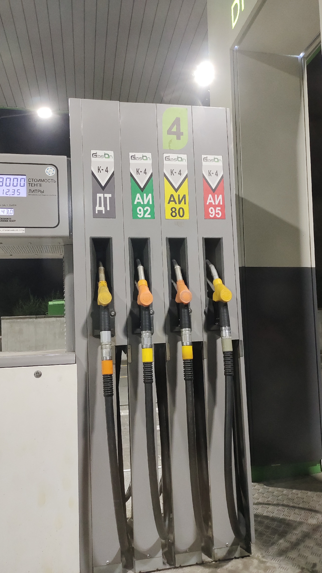 The image shows a close-up of a gas pump at a gas station. The pump is gray and has four different nozzles, each with a different colored handle: yellow, orange, yellow, and yellow. The nozzles are labeled with different types of fuel. The pump also has a digital display that shows the price and quantity of fuel. The background is blurred, but it appears to be a gas station with other pumps and a building. The lighting suggests it is nighttime or an indoor setting with artificial light.
