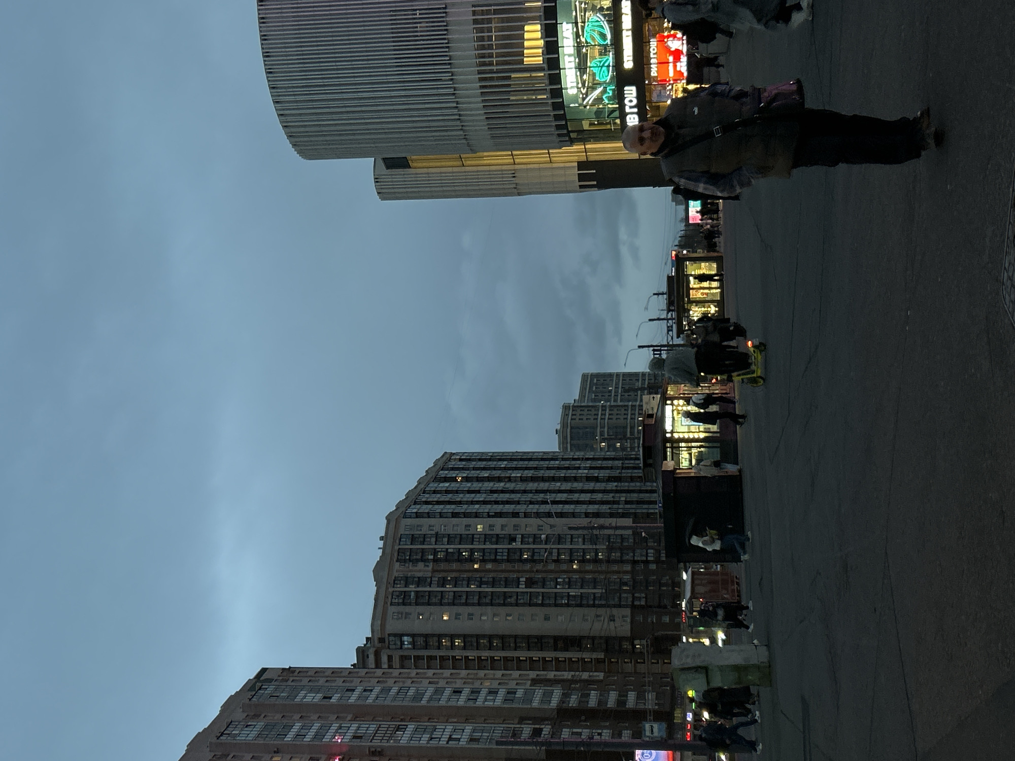 This media file depicts an urban street scene in Saint Petersburg, Russia, captured during dusk or early evening under an overcast, light blue-grey sky.
In the foreground, a paved street or square extends from the bottom of the frame. On the right, a man wearing a dark jacket, a plaid shirt, dark trousers, and carrying a cross-body bag, stands facing the viewer. Other individuals are visible further back on the pavement, walking or standing, suggesting general pedestrian activity.
The background and midground are filled with a variety of multi-story buildings. To the left, several large buildings with numerous windows, possibly residential or mixed-use, stretch upwards. On the right, a modern commercial building with distinctive horizontal architectural slats stands prominently. Below it, several brightly lit shopfronts and neon signs illuminate the scene. One clear sign features the English text "STREET BEAT" in green neon, accompanied by mirrored Cyrillic letters beneath it. The combination of fading natural light and glowing artificial lights creates a typical urban twilight atmosphere.