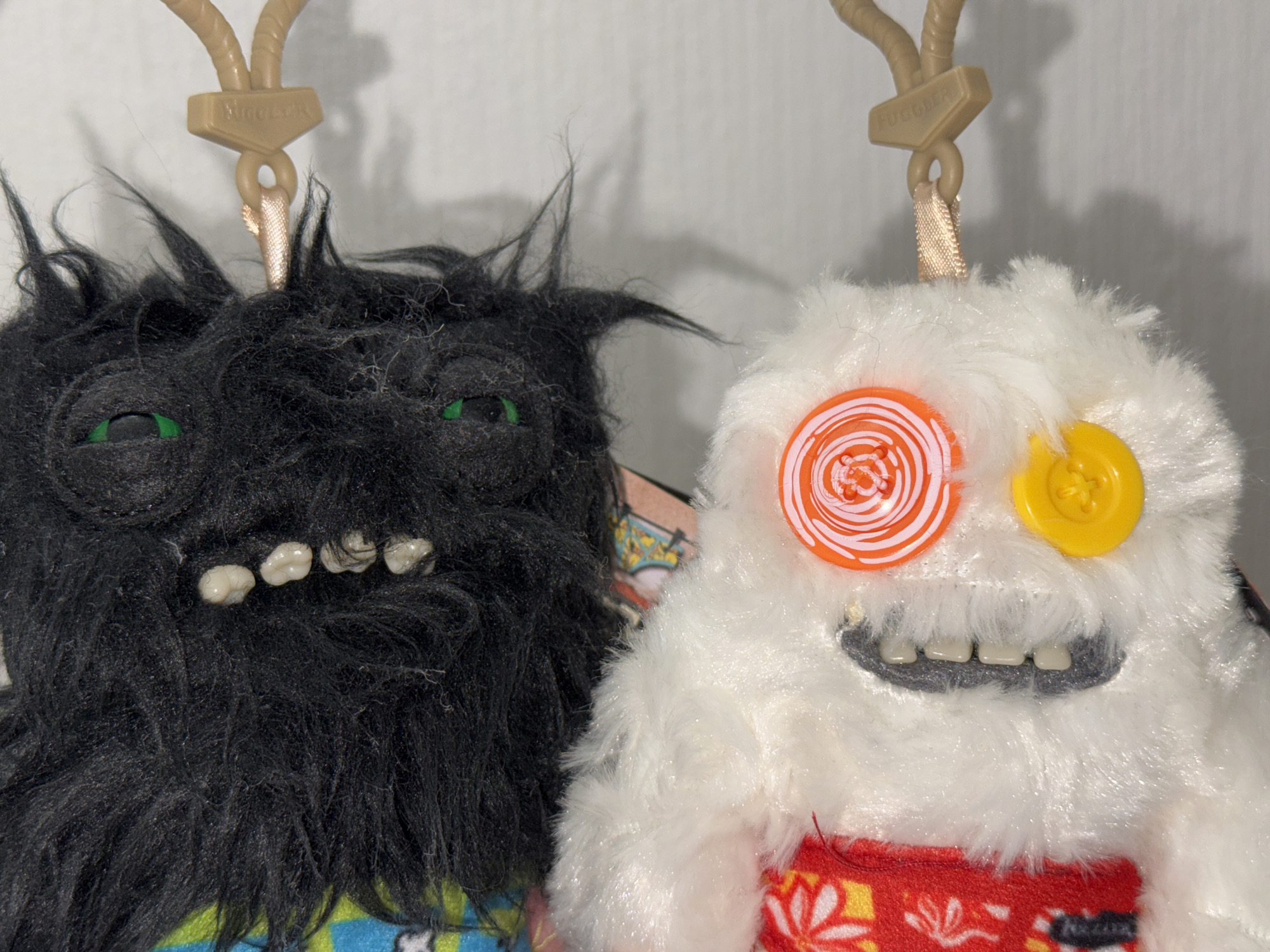 The image displays a close-up of two plush toy monsters, commonly known as "Fuggler" toys, positioned side-by-side against a plain, light-colored background.
The monster on the left is predominantly black, covered in shaggy, dark fur. It features two small, bright green eyes peeking through its fur and an open mouth revealing several uneven, off-white, human-like teeth. A tan plastic clip is visible at the top of its head, suggesting it is a keychain or can be hung.
The monster on the right is white, covered in fluffy, shaggy fur. Its eyes are mismatched buttons: one is a larger, round orange button with a white spiral pattern, and the other is a smaller, plain yellow button. It has a wide, smiling mouth showing a row of relatively uniform, off-white, human-like teeth. Below its mouth, a piece of red fabric with white and yellow floral or geometric patterns is visible, appearing to be clothing. A tan plastic clip, identical to the one on the black monster, is attached to the top of its head. The word "FUGGLER" is clearly embossed on this clip.
The toys appear to be in good condition, and the lighting is even, suggesting an indoor setting. There are no specific activities taking place, and the toys are static. No explicit time of day or location cues are visible within the image itself, though the provided context places the location in Saint Petersburg, Russia.