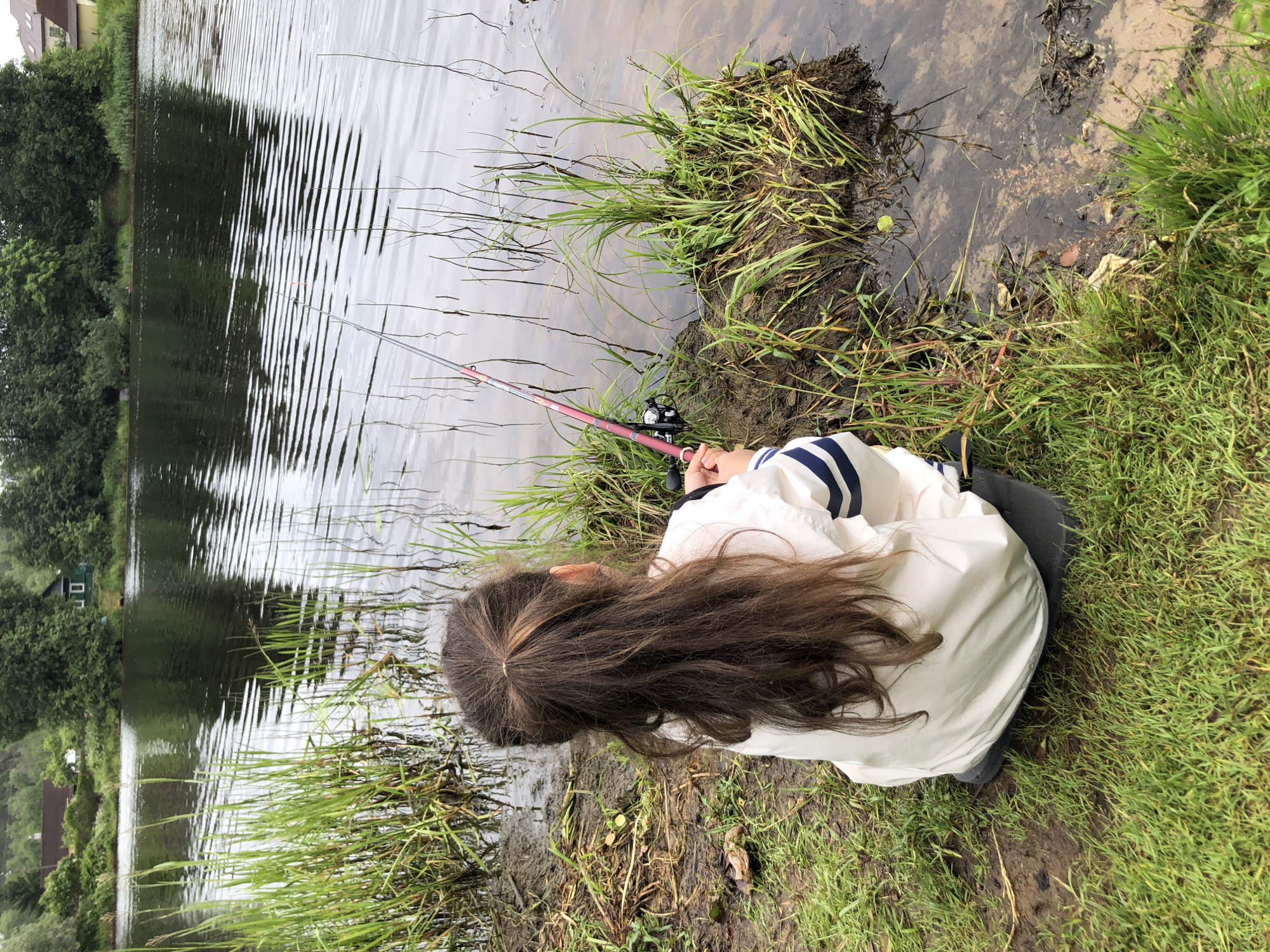 A young person with long brown hair is fishing on the edge of a body of water. They are sitting on the grassy bank, facing the water, and holding a fishing rod. The fishing rod is pink and silver with a black reel. The person is wearing a white jacket with blue stripes on the sleeves. The water is calm with ripples, and there is green grass and mud on the bank. In the background, there are trees and houses, suggesting a rural or semi-rural setting. The scene appears to be set during the daytime, under overcast or cloudy skies, as there are no harsh shadows. The overall atmosphere is peaceful and tranquil, with the focus on the quiet activity of fishing. There is no visible text in the image.