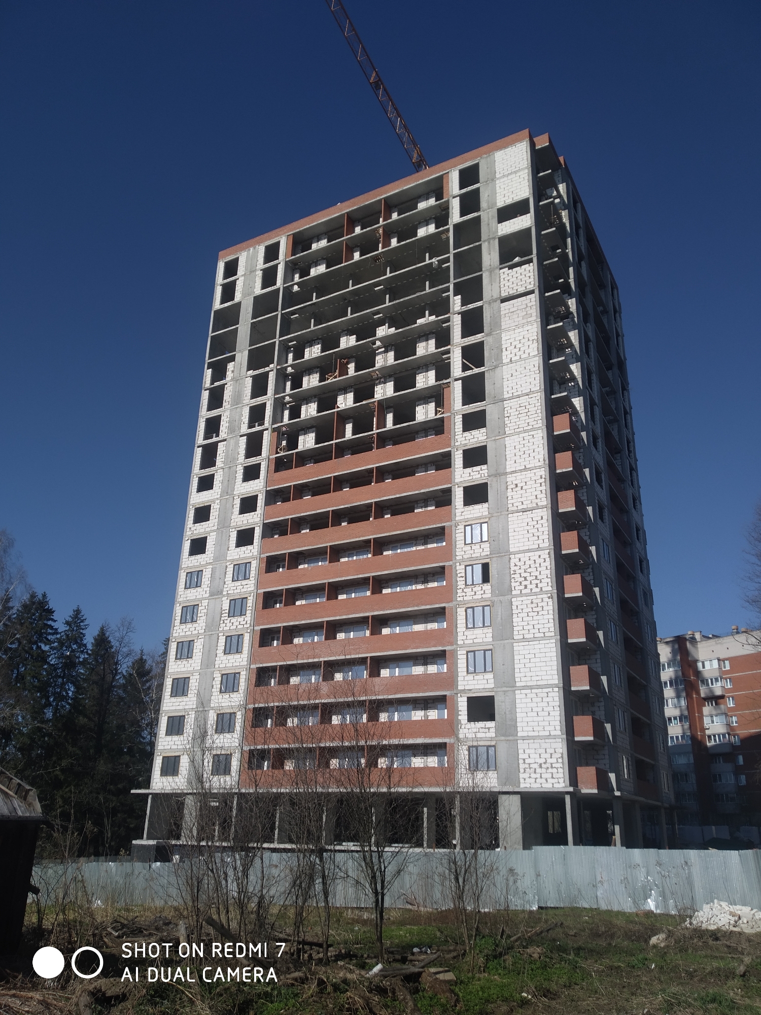 An upward-angle shot presents a partially constructed, multi-story building in Chaikovsky, Russia, under a cloudless blue sky. The building, likely residential, has a framework of concrete and white blocks with sections of red brick on several floors. Windows are installed in some areas. A construction crane is visible atop the building. The foreground contains low vegetation, bare tree branches, and a fence partially obscuring the building's base. Another finished residential building is visible in the background to the right of the construction site. A piece of equipment, likely a dumpster, is visible to the left of the frame. A watermark indicating the image was taken with a Redmi 7 AI Dual Camera is present at the bottom. The site appears to be in an early stage of construction or renovation.