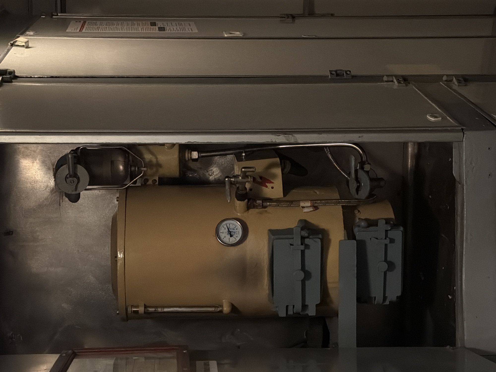 The image displays a detailed view of a complex piece of industrial or utility machinery, likely situated indoors within an equipment room or a large service unit. The central object is a substantial, horizontally oriented machine, predominantly tan or beige in color, which appears to be a motor, pump, or compressor.
Attached to this main machine are various components, including several metallic pipes and tubes that connect to different parts of the system, implying fluid or gas transfer. A notable detail on the side of the tan machine is a small, circular pressure gauge with a white face. The gauge displays measurement units in both "kgf/cm2" and "PSI," with numerical markings ranging from 0 to 160 PSI. The needle on the gauge indicates a pressure reading of approximately 40 PSI. Nearby, two gray, box-like components, each secured with a latch, are mounted on the side of the machine. To the far left, a smaller, dark, cylindrical part with a wheel-like attachment is visible, connected by a tube.
Above the main machinery, several layers of gray, metallic panels form an enclosure or housing, fastened by multiple latches. A rectangular label with unreadable text is visible on one of these top panels. A small red, arrow-shaped symbol is also seen near a valve on the tan machine. The scene is devoid of people and appears to be illuminated by artificial light, characteristic of an industrial or mechanical setting. The context suggests this equipment is located in Kirsanov, Russia.