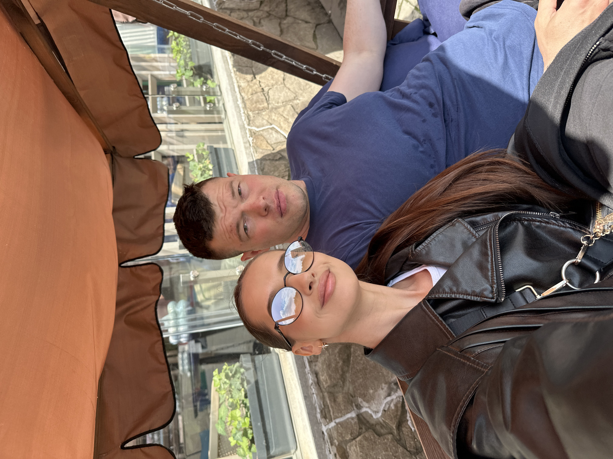 A man and a woman are posing for a selfie on a swing set under a brown awning. The woman is in the foreground, wearing a black leather jacket and round sunglasses. She is smiling and has her hair down. The man is behind her, wearing a blue t-shirt. They appear to be at an outdoor cafe or restaurant. The background shows a stone wall and a window with plants. The image was taken in the city of Yekaterinburg, Russia.