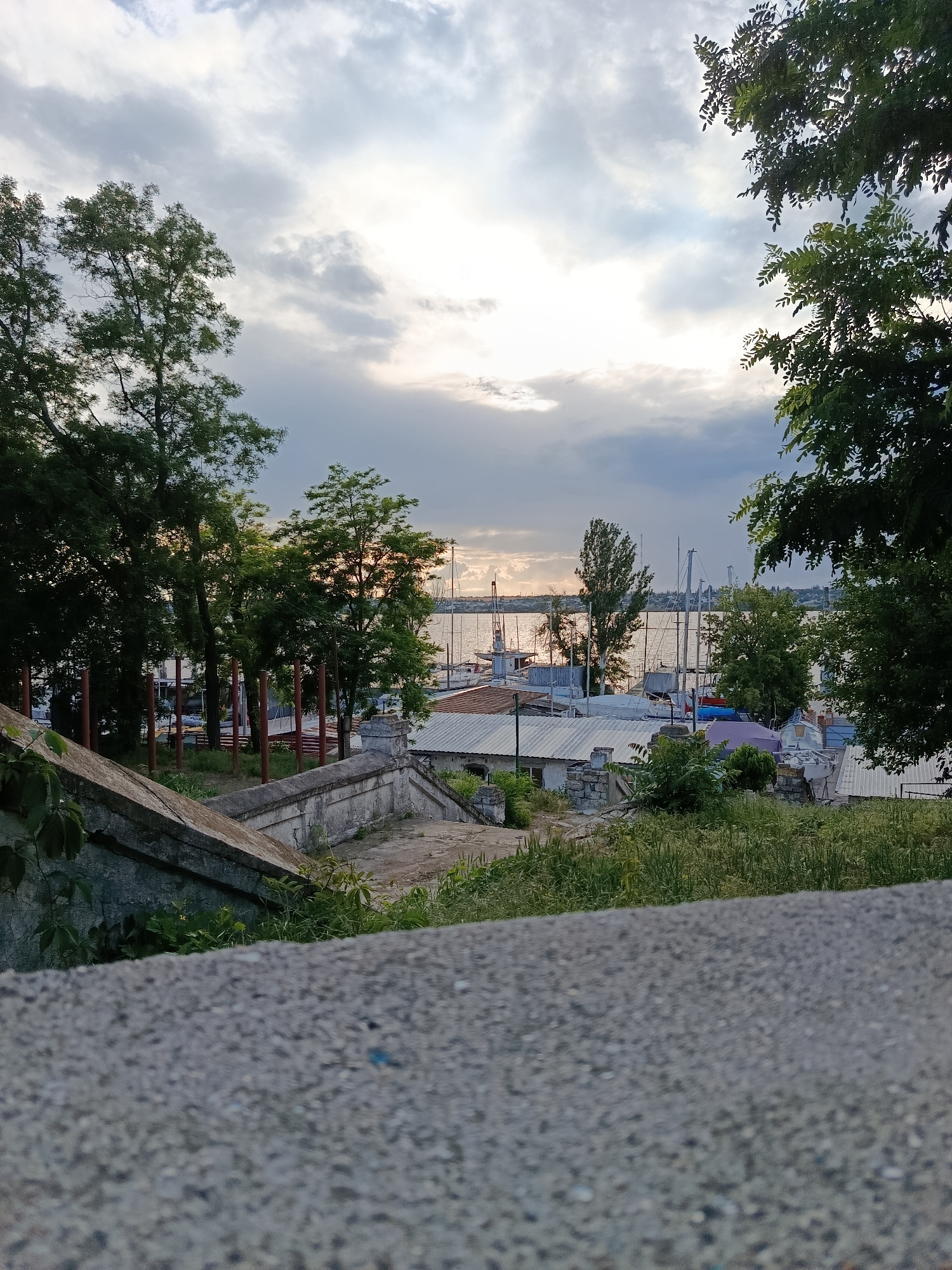 A high-angle shot captures a hazy, overcast sky with sunlight peeking through the clouds, casting a soft glow on a waterfront scene. The foreground is dominated by a rough, grey concrete surface, creating a shallow depth of field that blurs the details. Beyond this, a worn, concrete staircase descends towards a grassy embankment.
Further down, a cluster of trees with lush green foliage frames the middle ground. Between the trees, a harbor is visible, filled with various sailboats, their masts reaching towards the sky. Some boats are covered, while others are partially obscured. A few low-lying buildings with corrugated metal roofs are scattered near the waterfront. The distant horizon shows a faint outline of land. No people are visible in the image. The overall mood is serene and slightly melancholic, with the diffused light and muted colors.