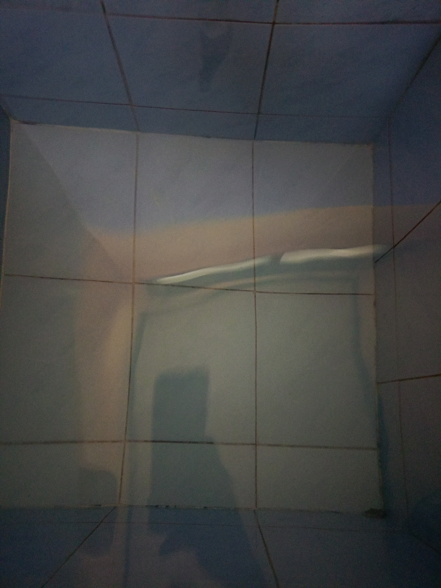 The image shows a corner of a tiled room, possibly a bathroom. The tiles are light blue and off-white. Grout lines are visible between the tiles. There are shadows and reflections on the tiles, including a shadow resembling a person holding a phone. The overall lighting is dim. It is not possible to determine the location in Bireuen, Indonesia from the image.