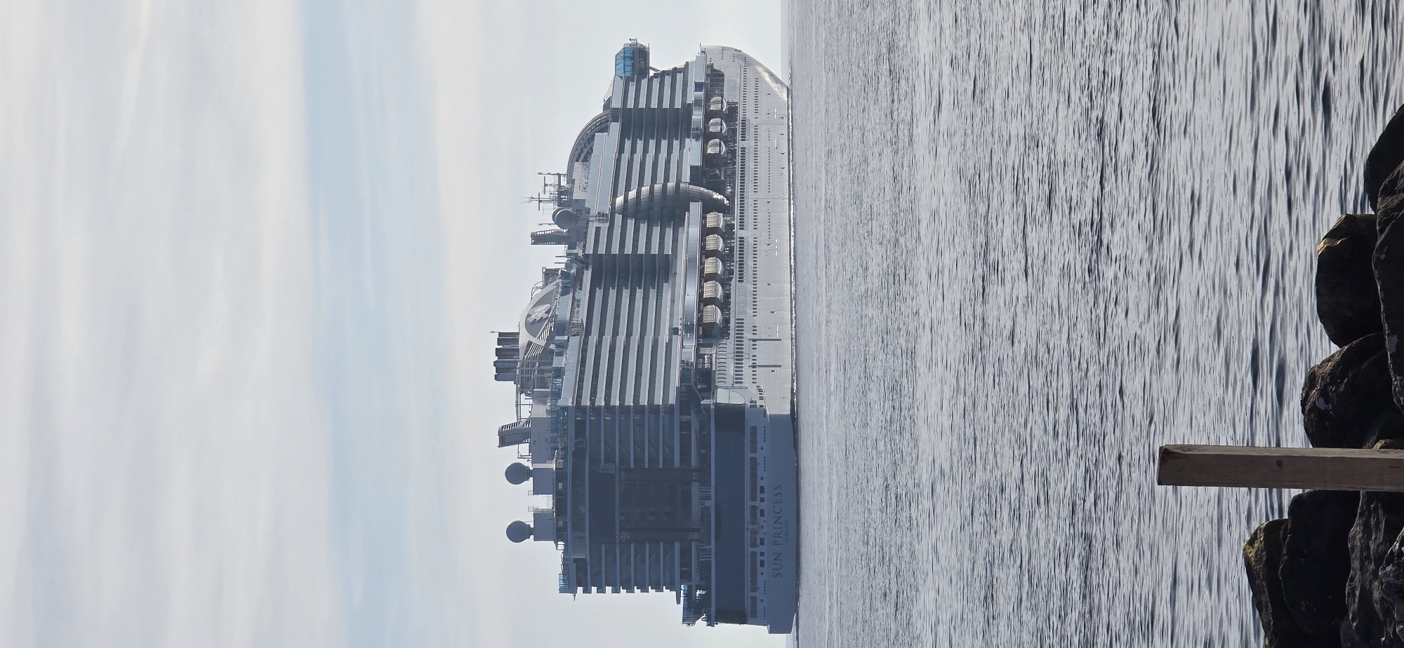 A large cruise ship, identified by the visible text "SUN PRINCESS" on its hull, navigates calm, rippled water under a bright, overcast sky. The ship, a prominent subject on the left and center, features multiple decks with numerous balconies and windows, rendered in shades of white and grey. Distinctive blue-tinted glass structures and various communication antennae are visible on its upper levels. Lifeboats are neatly lined along the ship's side.
On the right side of the frame, a rugged shoreline with dark, textured rocks provides a natural contrast to the open water. A simple wooden plank extends horizontally from these rocks, appearing to jut out over the water. The sky is a soft blend of light blue and white, indicative of daytime with diffused sunlight. The scene suggests a tranquil moment in a coastal setting, consistent with the provided location context of Bar, Montenegro. There are no people visible.