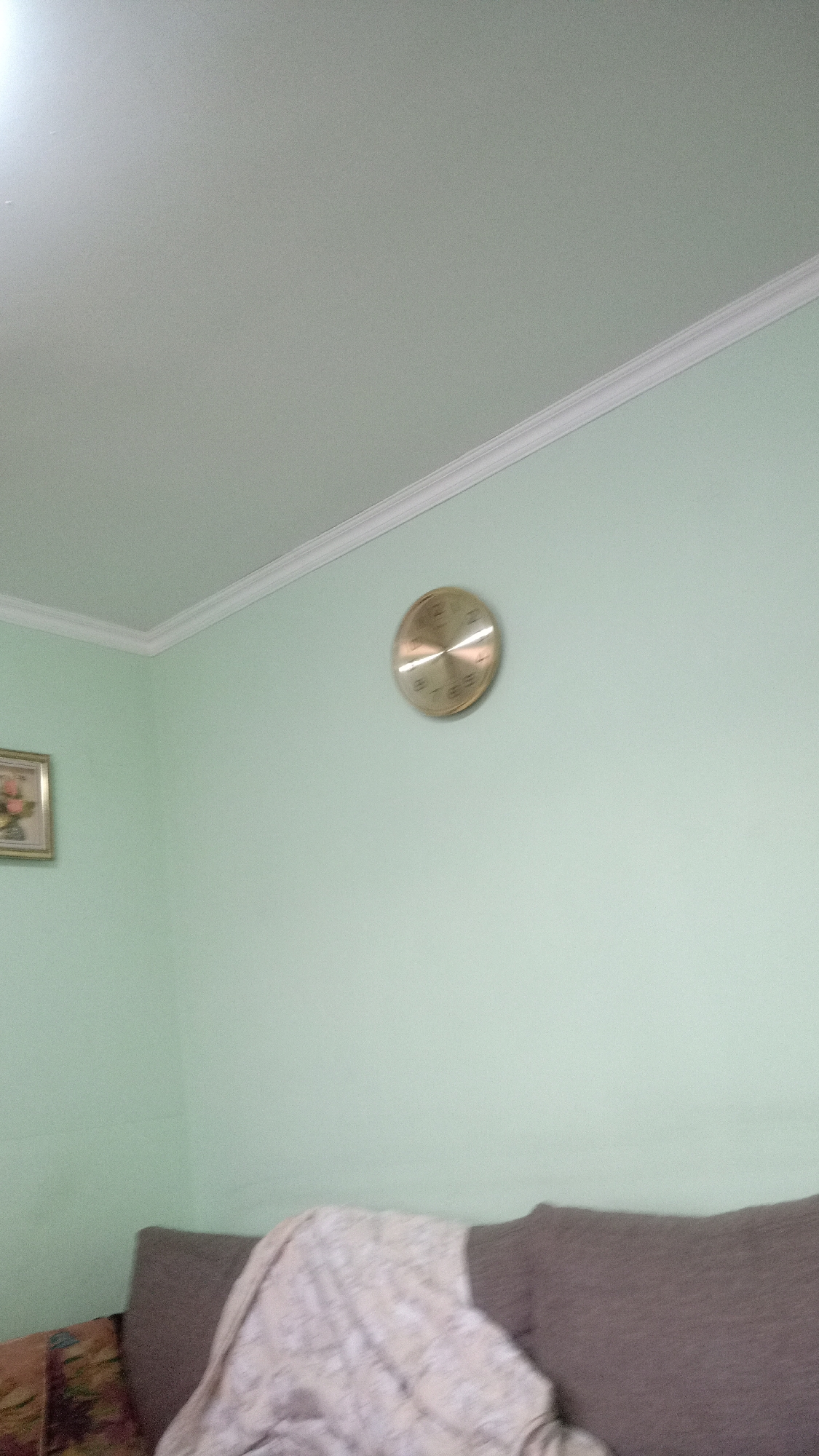 The image shows an interior view of a room. The walls are painted a light green color with white trim along the ceiling. A round, gold-colored clock is mounted on the right wall. To the lower left, there is a framed picture hanging on the wall. In the foreground, the top portion of a couch is visible, draped with a light-colored patterned blanket.