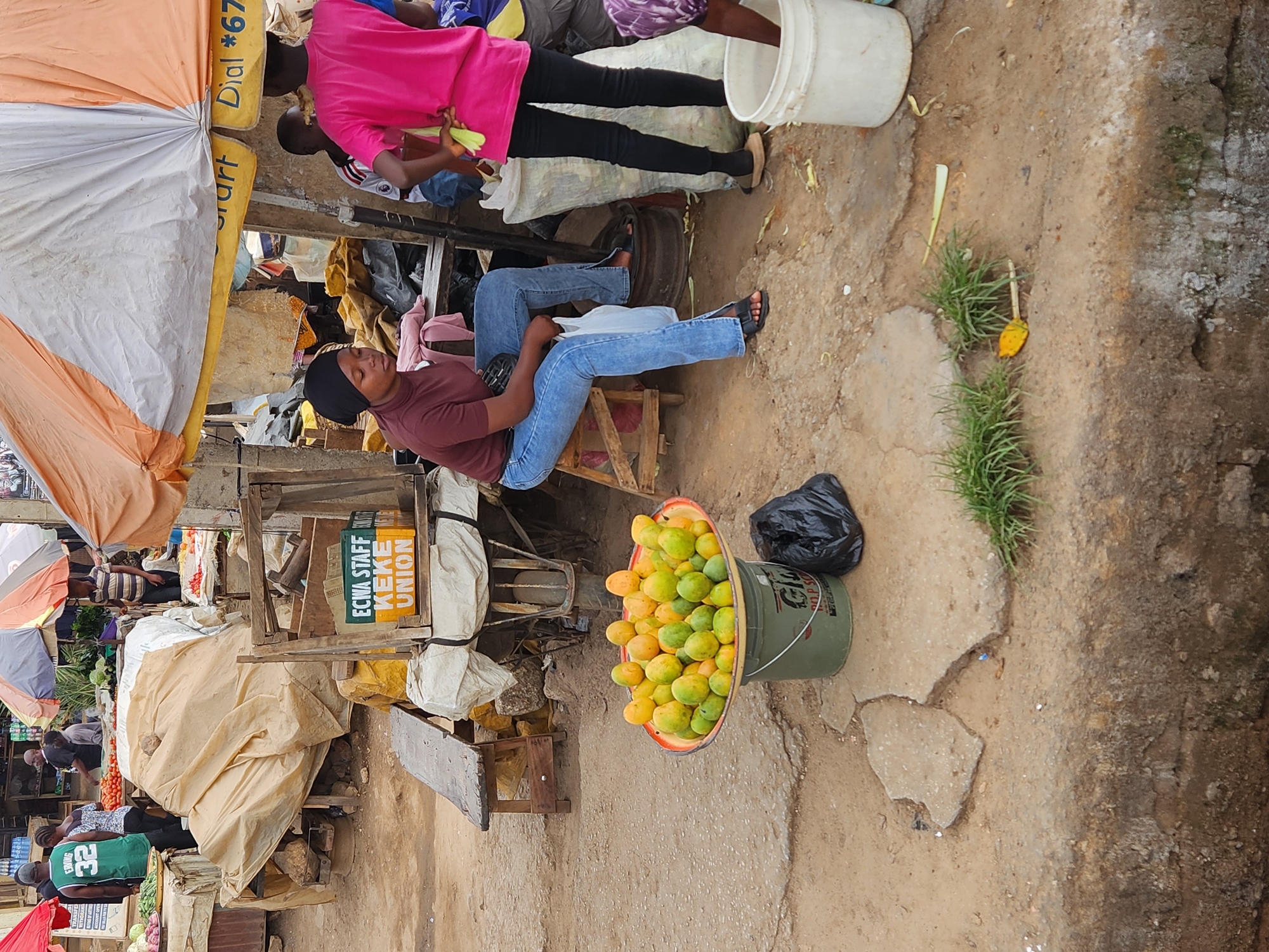 The image depicts a street scene in Jos, Nigeria. A woman wearing a maroon shirt, blue jeans, and a black head covering is seated. Next to her is a bucket overflowing with mangoes and a black plastic bag. Behind her is a stall with various items under a colorful umbrella. Other people are visible in the background, including one wearing a pink shirt. The ground is a mix of dirt and concrete.