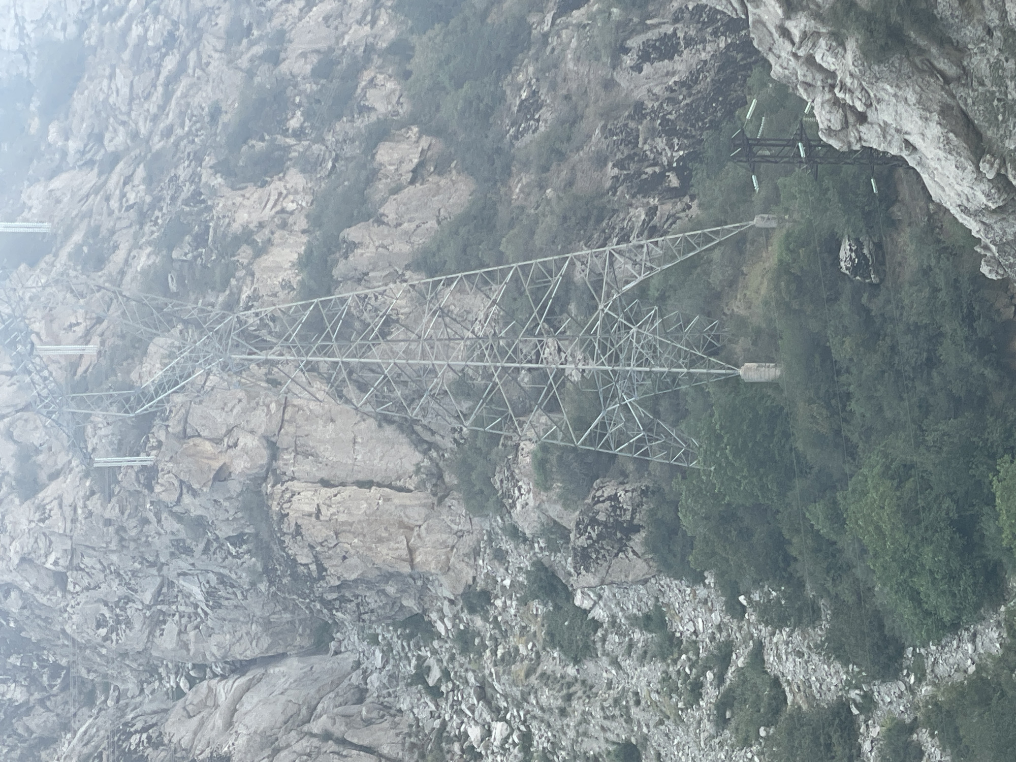 The image captures a steep, rocky mountainside in Khodzha-Obigarm, Tajikistan, dominated by towering electrical transmission pylons. The terrain is rugged, with loose rocks and sparse vegetation clinging to the slopes. Several large, metallic pylons are visible, some with intricate lattice structures, supporting power lines that traverse the landscape. One pylon is particularly prominent, extending high up the mountain. Below the larger pylons, a smaller, darker pylon with insulators is also present, positioned amidst dense green foliage. The overall scene conveys a sense of industrial infrastructure integrated into a natural, mountainous environment, with the haze suggesting distance or atmospheric conditions.