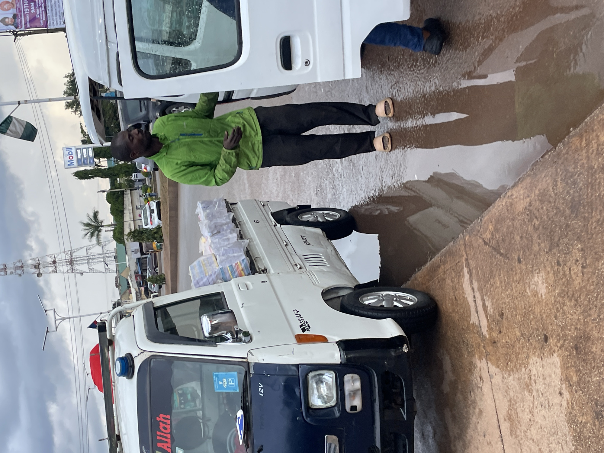 The image depicts a street scene in Jos, Nigeria, under an overcast sky. In the foreground, a white and dark blue kei truck is parked, loaded with stacked items wrapped in plastic. A man wearing a green jacket and black pants, with light-colored slip-on sandals, stands next to the truck, his hand on the open door of a white van. Another person, only their legs and feet wearing dark shoes and blue jeans are visible, is partially in the van. The wet ground suggests recent rain, with puddles reflecting the surroundings. In the background, a tall communications tower, a Mobil gas station sign, and other buildings are visible. The overall atmosphere suggests a bustling urban environment, possibly a market or transportation hub, with ongoing activity despite the weather.