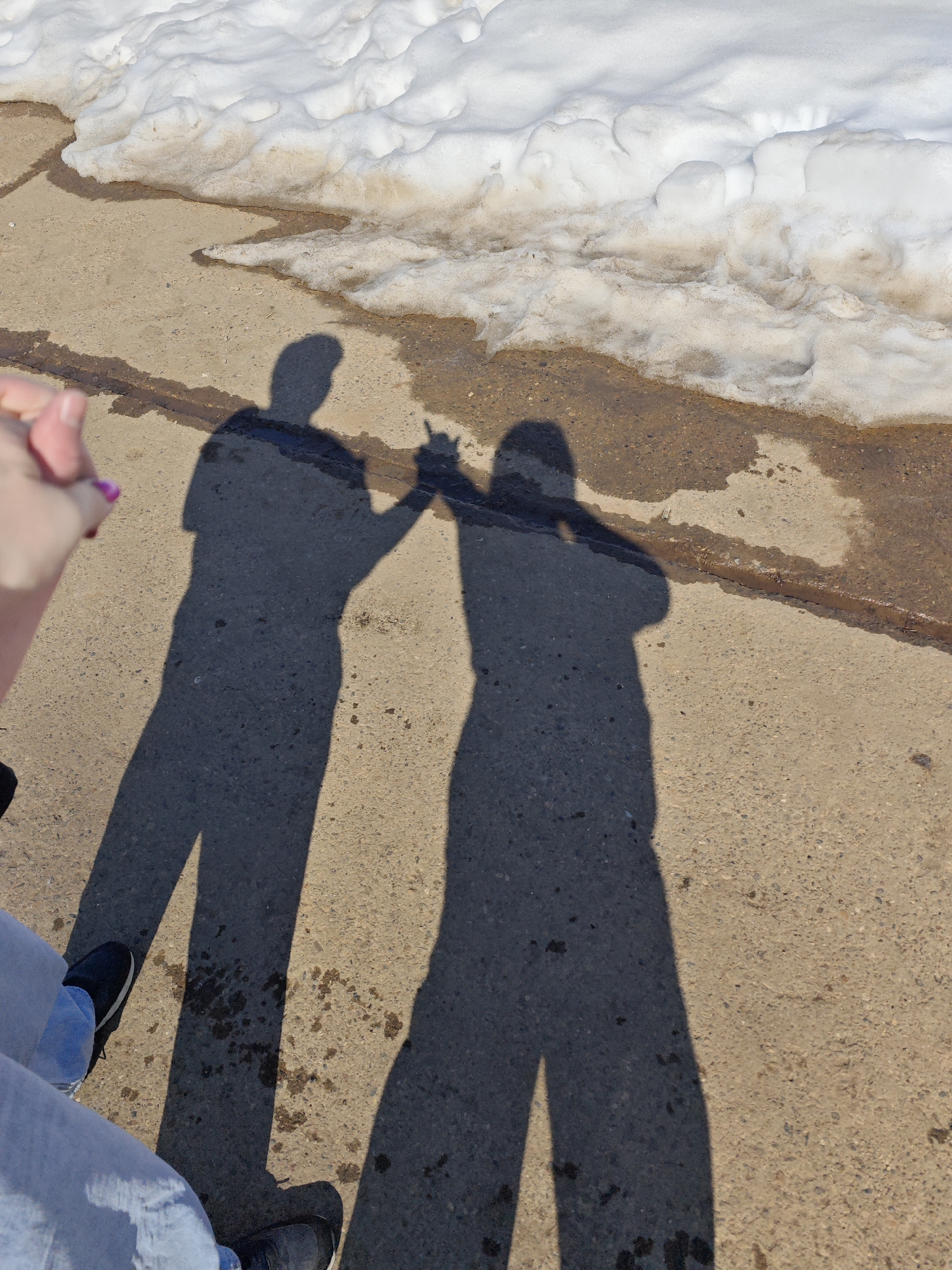The image depicts a concrete sidewalk with patches of snow melting along the edge. Two shadows are cast on the sidewalk, suggesting two people interacting with each other. One shadow appears to show a person holding up a single finger. A person is seen in the bottom left of the image, wearing blue jeans and black shoes. The location is suggested to be Марковский, Russia.