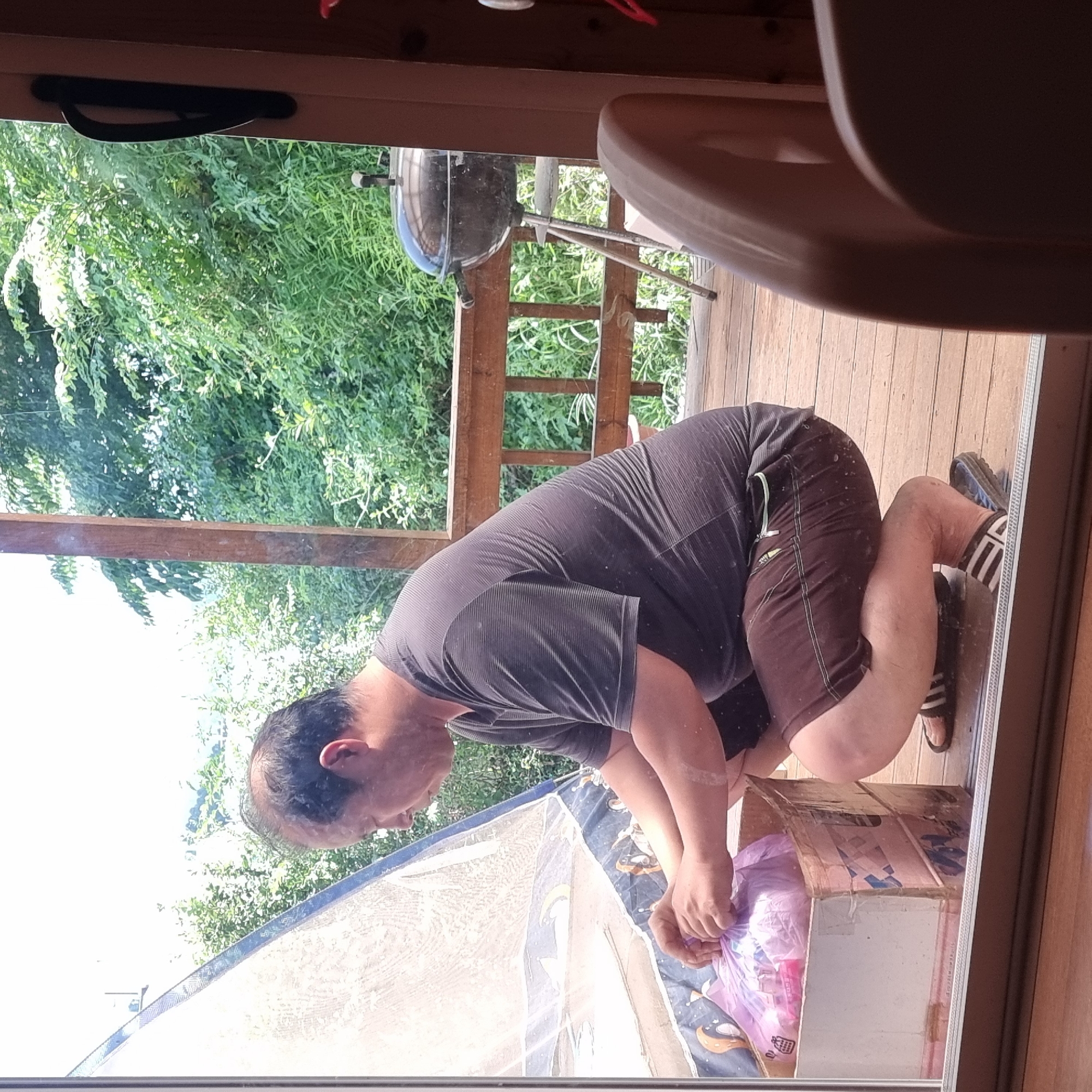 In Dunnae-myeon, South Korea, a man is seen crouching on a wooden deck next to a sliding glass door. He is wearing a dark, short-sleeved, striped t-shirt and brown shorts with a green zipper detail. He is holding a pink plastic bag filled with items and appears to be placing it into a cardboard box. A grill is visible on the deck behind him, and lush green foliage surrounds the area. The scene suggests a domestic or outdoor living space.