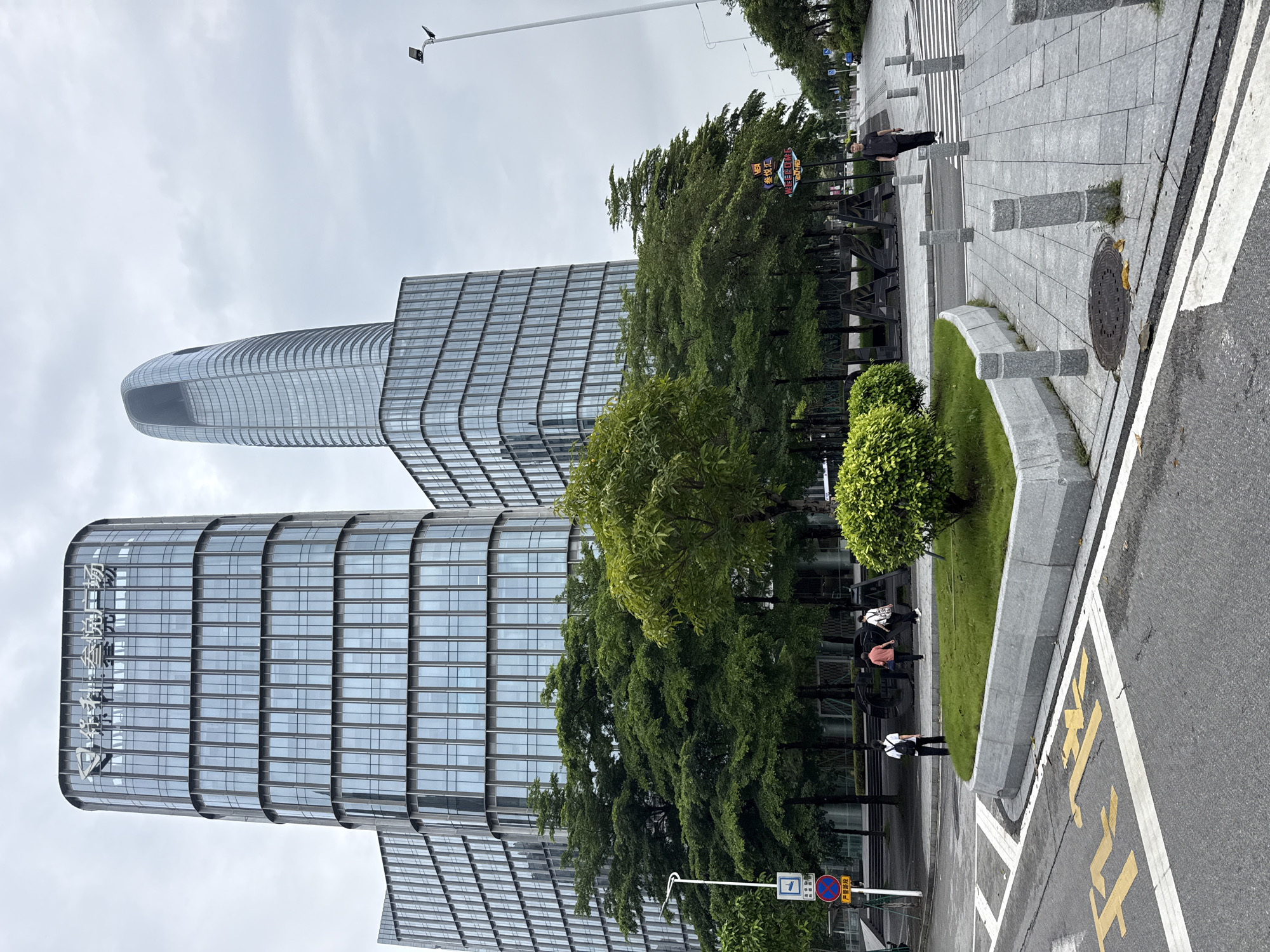 This is a vertical, eye-level, medium shot of a modern street scene in Haizhu District, China. The main subjects are two contemporary, glass-paneled office buildings with curved facades. One building is taller and features a rounded, elongated top, while the other is shorter and wider. Green trees and manicured bushes line the sidewalk, creating a sharp contrast with the metallic and glass structures.
Several individuals are visible on the sidewalk. One person in dark clothing walks along the paved area, while a group of three people, one in an orange shirt and two in darker attire, are near a black, abstract sculpture. Further along the sidewalk, another person in dark clothing with a backpack walks. The ground is paved with large, light-gray tiles, and a crosswalk with white stripes is present. A circular manhole cover with some debris on it is visible near the curb. A yellow painted marking, possibly indicating a parking space, is on the asphalt road.
A street lamp with a small camera attached is visible in the upper portion of the image, suggesting daylight. The sky is overcast and white, indicating a cloudy day. A street sign with Chinese characters is visible near the trees. The overall mood is calm and orderly, typical of an urban business district.