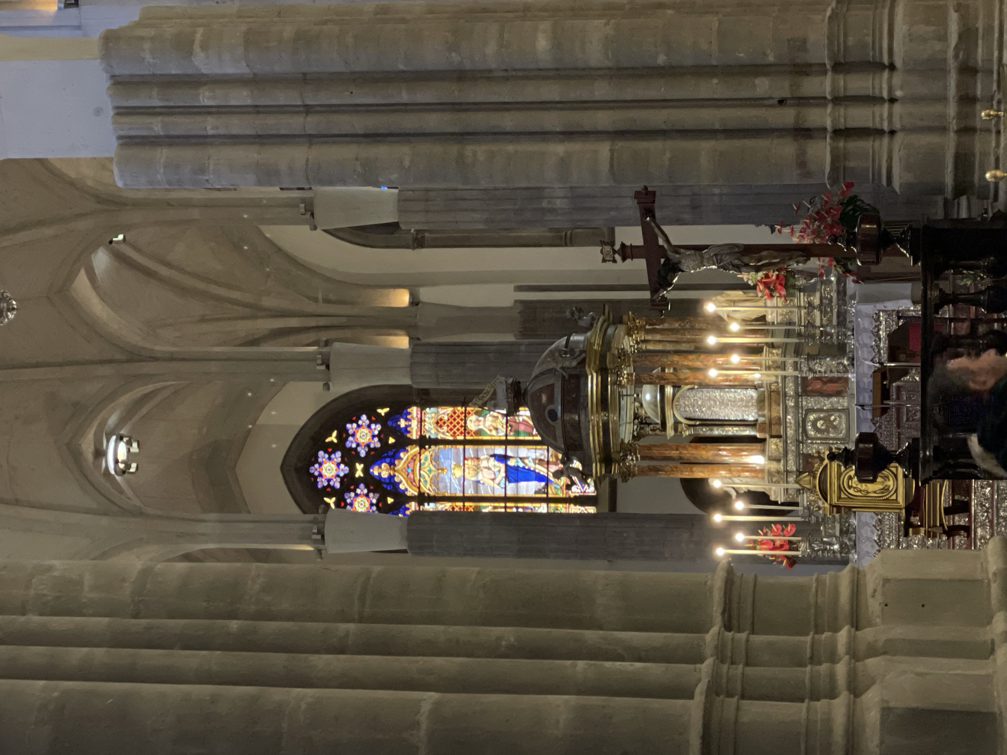 The image displays the interior of a grand religious building, likely a church or cathedral, in San Cristóbal De La Laguna, Spain. Massive, fluted stone columns rise vertically, supporting vaulted ceilings and intricate architectural details. The overall setting is characterized by a solemn and historic atmosphere.
The central focus is an ornate altar area in the mid-ground. A prominent wooden crucifix, depicting the figure of Jesus Christ, is positioned above this structure. Below it, a richly decorated shrine or tabernacle gleams with golden and silver elements, adorned with intricate patterns and reflective surfaces. Several lit candles in tall candelabras cast a warm glow, interspersed with red floral decorations. Behind this, large, reflective panels with embossed or engraved designs appear to be made of silver or polished metal.
To the left of the altar, a vibrant stained-glass window stands out, illuminating the space with a burst of color. The window features religious figures, including a central figure predominantly in blue and yellow, surrounded by elaborate circular and star-like patterns in hues of red, purple, and green.
The lighting within the space is dim, augmented by the glow of the candles and various artificial light fixtures on the vaulted arches, suggesting either evening or a subdued interior environment. A partial view of a person's head and shoulder is visible on the far right, near the altar. No discernible text is visible in the image.