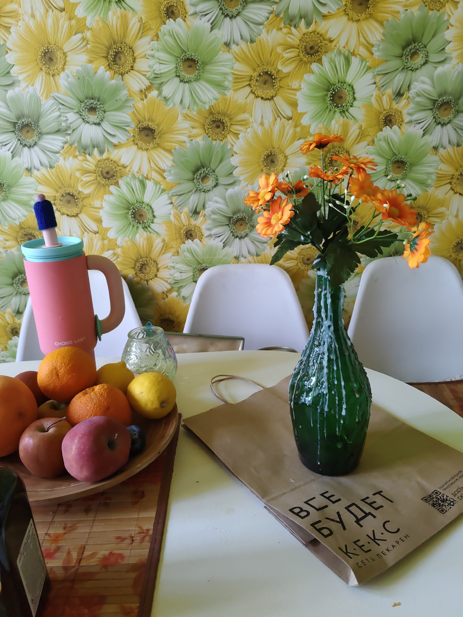 A still life photograph captures a collection of items on a white table against a backdrop of floral wallpaper. In the foreground, a wooden bowl holds an assortment of fruits, including oranges, lemons, apples, and what appears to be a plum. To the left of the bowl, a pink travel mug with a light blue lid and a pink handle is positioned. Behind the fruit bowl and mug, a green glass bottle serves as a vase for a bouquet of artificial orange flowers, with white wax dripping down the sides of the bottle. A small, clear glass jar with a decorative lid sits between the mug and the fruit bowl. To the right of the vase, a brown paper bag with Cyrillic text and a QR code is visible. Two white chairs are partially visible in the background, and the wallpaper features a repetitive pattern of yellow and green daisy-like flowers. The lighting suggests an indoor setting, likely during the daytime. The provided location of Rostov-on-Don, Russia, is a detail that cannot be independently verified from the visual information in the image alone.