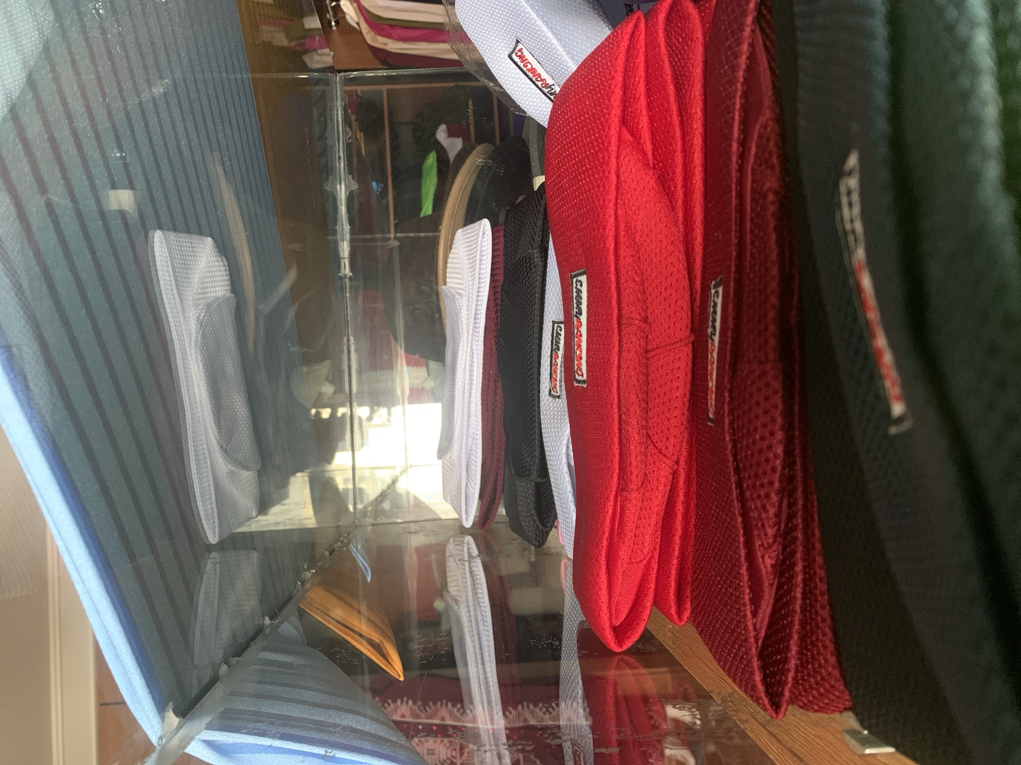 This image depicts a retail display of prayer caps, likely located within a shop in the city of Gura Top, Nigeria. The caps are stacked and arranged on shelves within a glass-fronted display case. The foreground features several layers of folded prayer caps in vibrant red, black, and white. Some of the caps have embroidered labels visible, one clearly showing the word "CAMPURANKING". Behind these, more caps in white and maroon are visible. The background reveals other items for sale, including clothing on hangers, and the interior of the shop, suggesting a market or retail environment. The lighting appears to be artificial, illuminating the merchandise within the display case.
