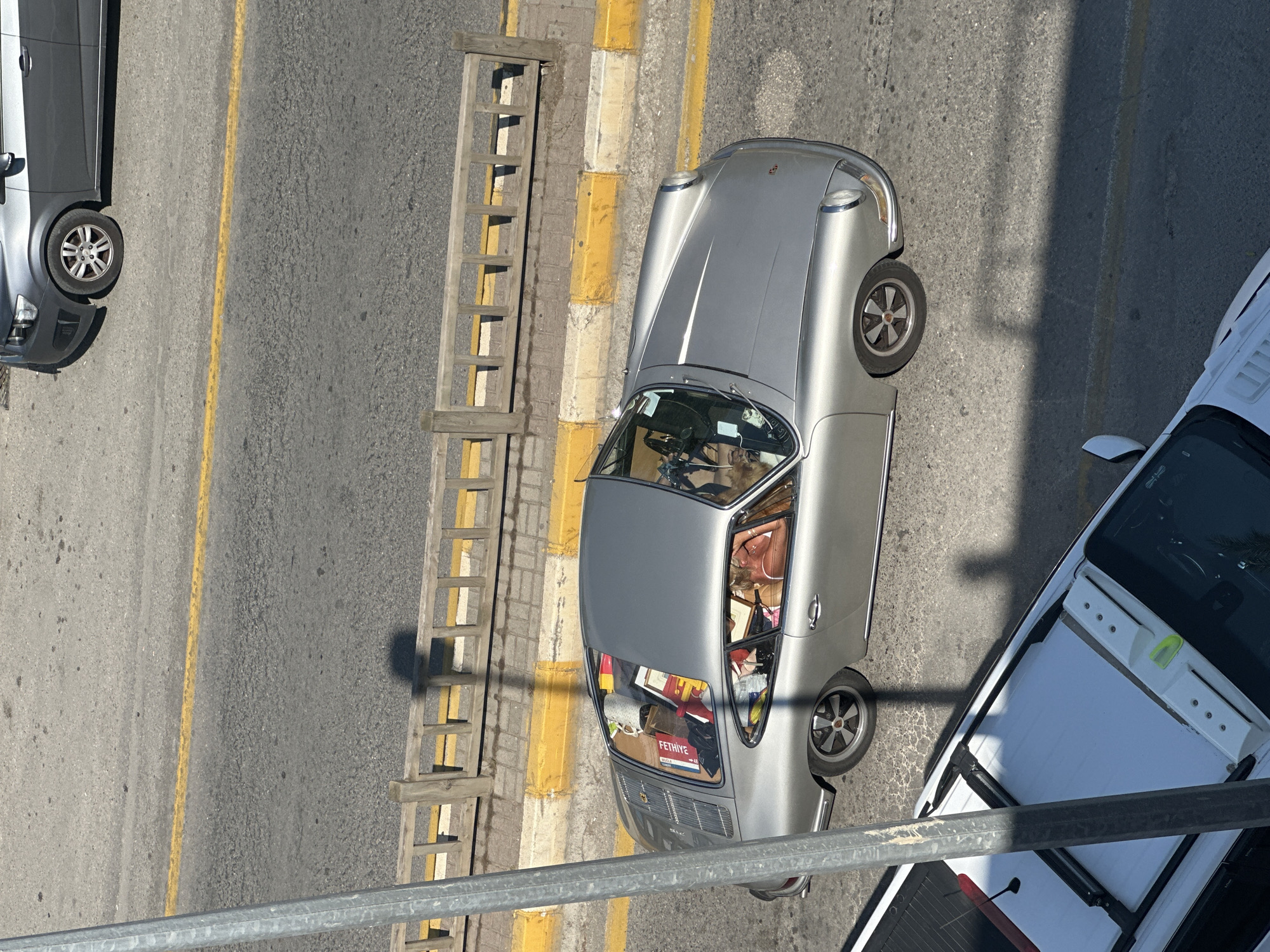 A silver classic Porsche coupe is parked on a street in Fethiye, Turkey. The car is facing away from the viewer, and there is a woman lying in the passenger seat with her head near the windshield. The back of the car is filled with luggage and boxes, one of which has "FETHIYE" printed on it. The car is parked alongside a curb with alternating yellow and white painted sections, next to a wooden ladder-like structure. To the left of the Porsche, partially visible, is a dark gray car. To the right of the Porsche, also partially visible, is a white pick-up truck with a covered bed. The scene is viewed from an elevated position, possibly from a building or bridge, and there are shadows cast across the street. A metal railing is in the foreground, suggesting the image was taken from a balcony or similar structure. The asphalt street has a yellow line painted on it.