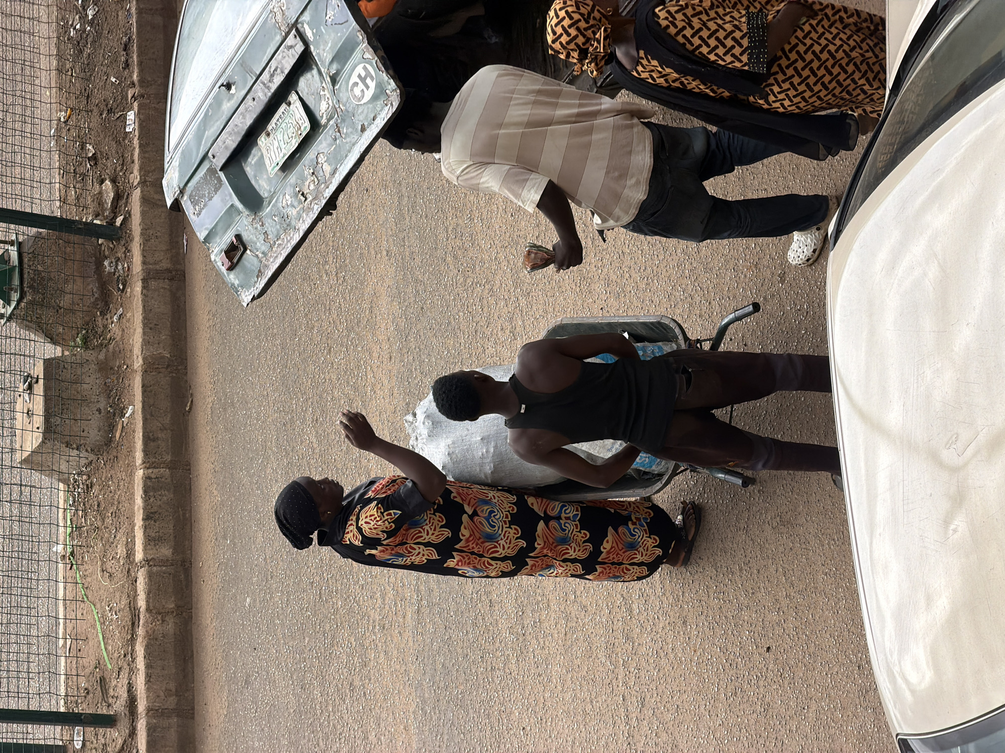 In Bukuru, Nigeria, on a paved street, four people are gathered around a wheelbarrow and the open trunk of a vehicle. A woman wearing a black headscarf and a vibrantly patterned dress stands to the left, holding something in her hand. To her right, a person in a black tank top and brown pants pushes the wheelbarrow, which contains a large white bag. Facing them, a man in a striped t-shirt and jeans holds a small amount of cash. To the far right, a woman in an orange and black patterned dress stands beside the open trunk of a weathered green vehicle. The trunk is propped open, revealing a license plate that reads "PLATEAU RY 2394 CH." The ground is a mixture of asphalt and loose gravel. A chain-link fence and concrete barrier line the edge of the road. A car hood is visible in the foreground.